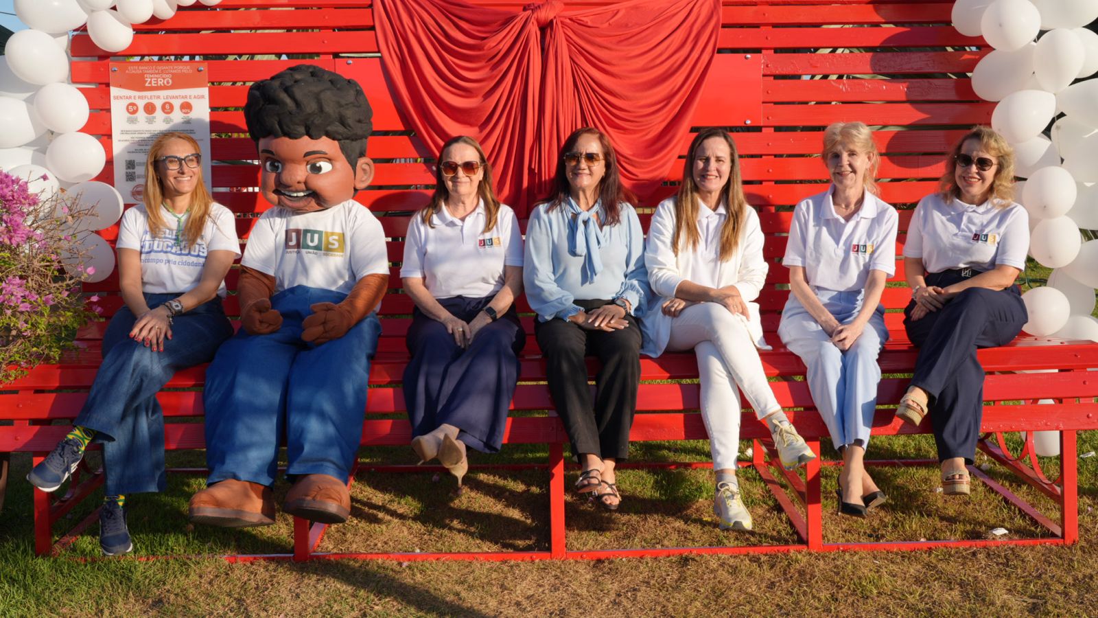A imagem mostra seis mulheres e um personagem fantasiado sentados em um grande banco vermelho. Ao fundo, há uma estrutura de madeira vermelha decorada com um tecido vermelho e balões brancos. O grupo posa para a foto durante uma ação do projeto Banco Vermelho.