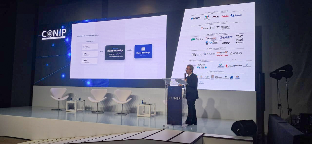 A imagem mostra o palco principal do 21º CONIP – Congresso de Inovação no Poder Judiciário e Controle. À direita, um homem de terno escuro faz uma apresentação em um púlpito transparente com a marca do CONIP. Ao fundo, dois grandes telões ocupam a parede: o da esquerda exibe um fluxograma do novo Sistema de Diário da Justiça, com caixas azuis conectadas por setas; o da direita reúne os logotipos de patrocinadores e apoiadores do evento. À esquerda do palco, há três poltronas brancas e pequenas mesas de apoio com garrafas de água.