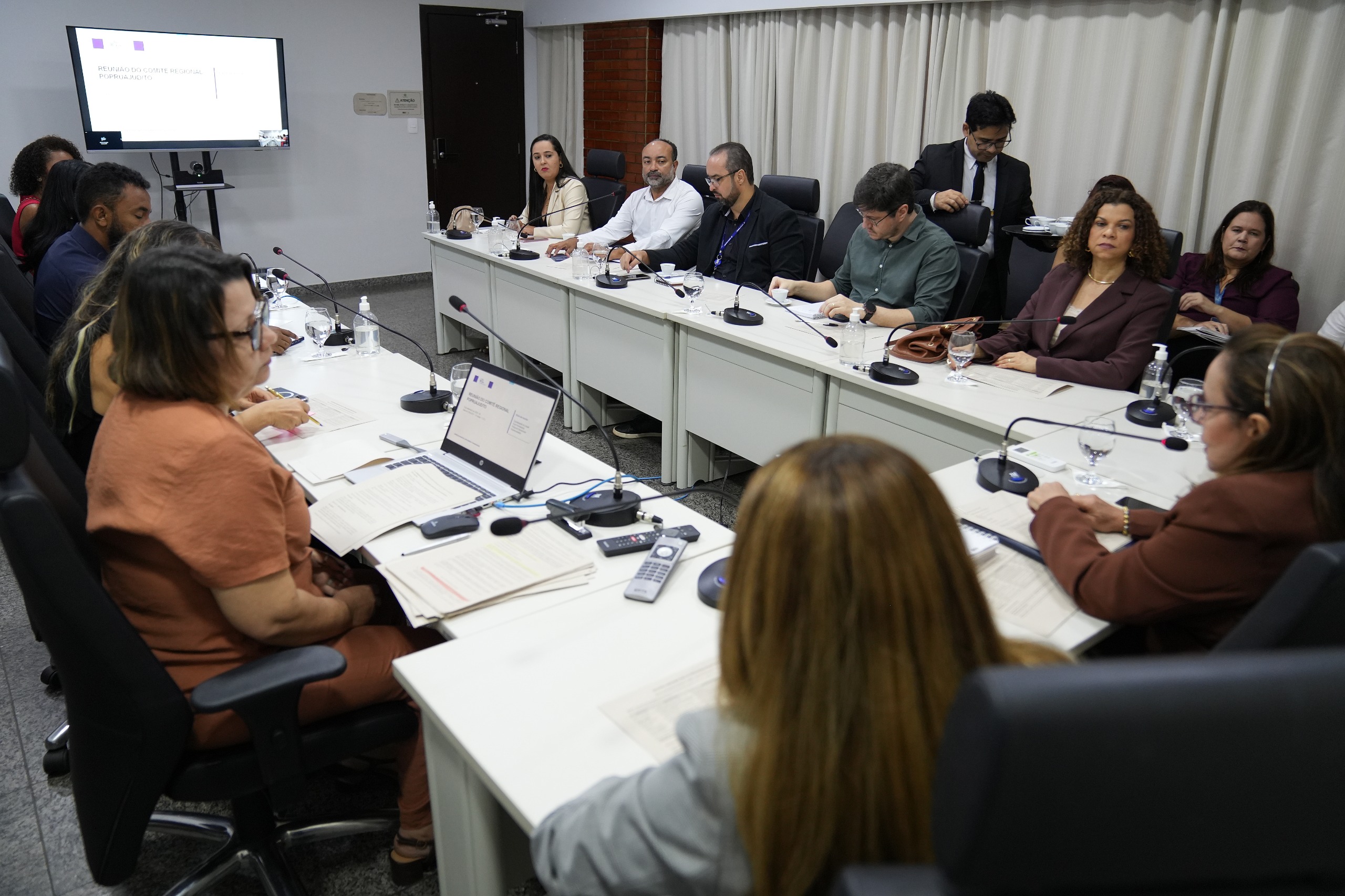 Representantes de diferentes instituições participam da reunião do Comitê Regional PopRuaJud/TO, no Tribunal de Justiça do Tocantins. Sentados ao redor de mesas, os participantes acompanham as discussões com documentos, computadores e microfones. Ao fundo, uma tela exibe a identificação da reunião.
