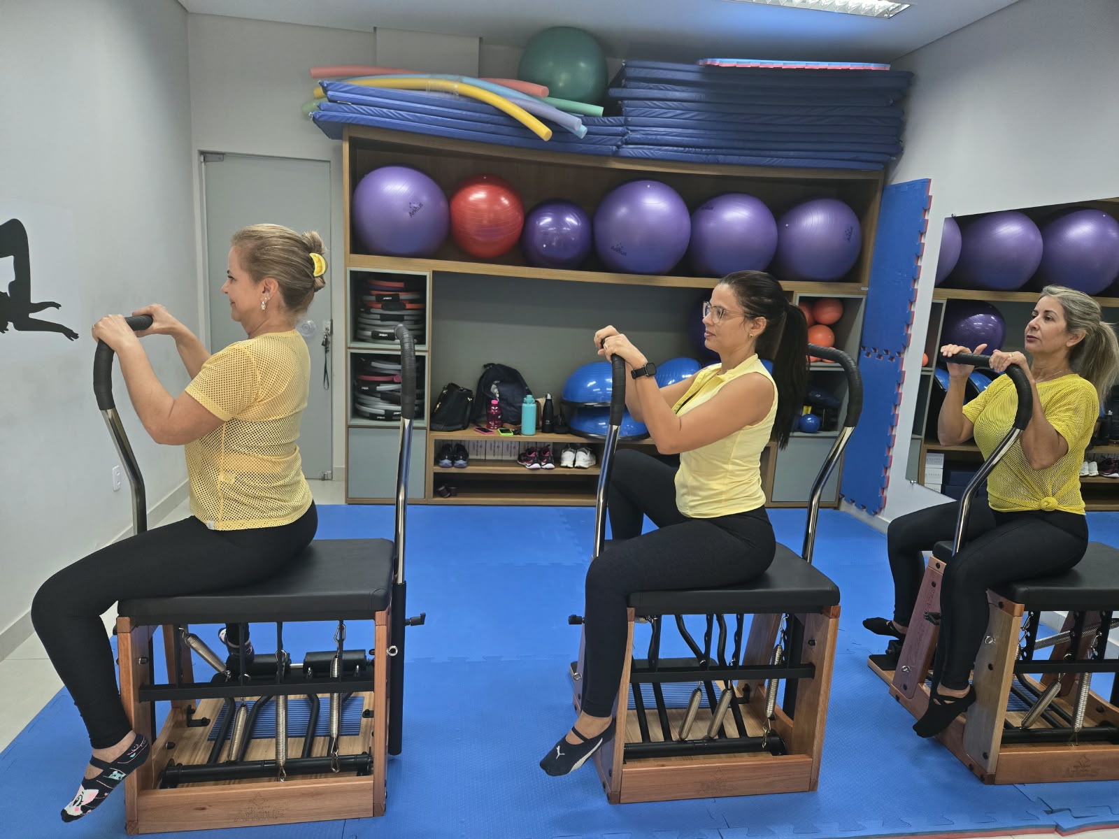 Três participantes, vestidas de amarelo, fazem um exercício de Pilates sentadas em equipamentos, com as mãos apoiadas nas barras à frente do corpo. Ao fundo, prateleiras guardam bolas, colchonetes e outros materiais usados nas aulas.