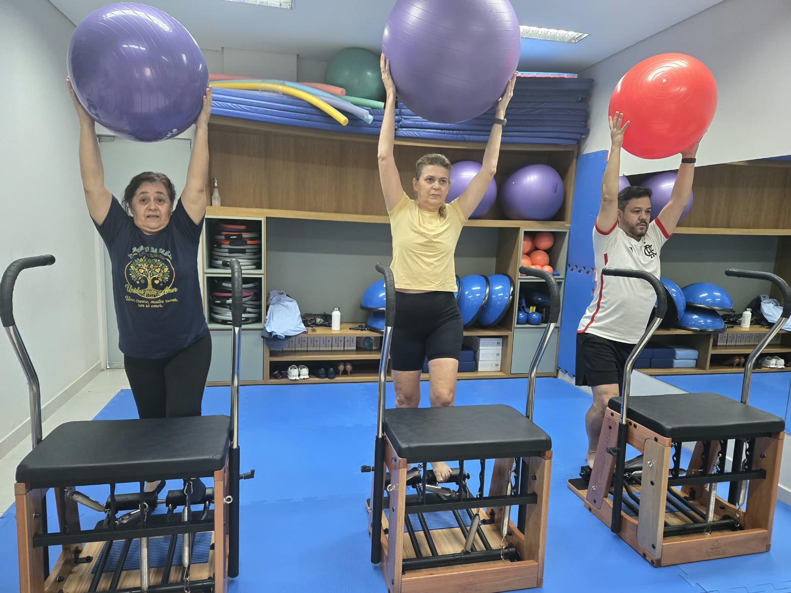 Três participantes de uma aula de Pilates Terapêutico levantam bolas de exercício acima da cabeça. À frente de cada um há um equipamento de Pilates; ao fundo, prateleiras guardam colchonetes e outros materiais usados nas atividades.