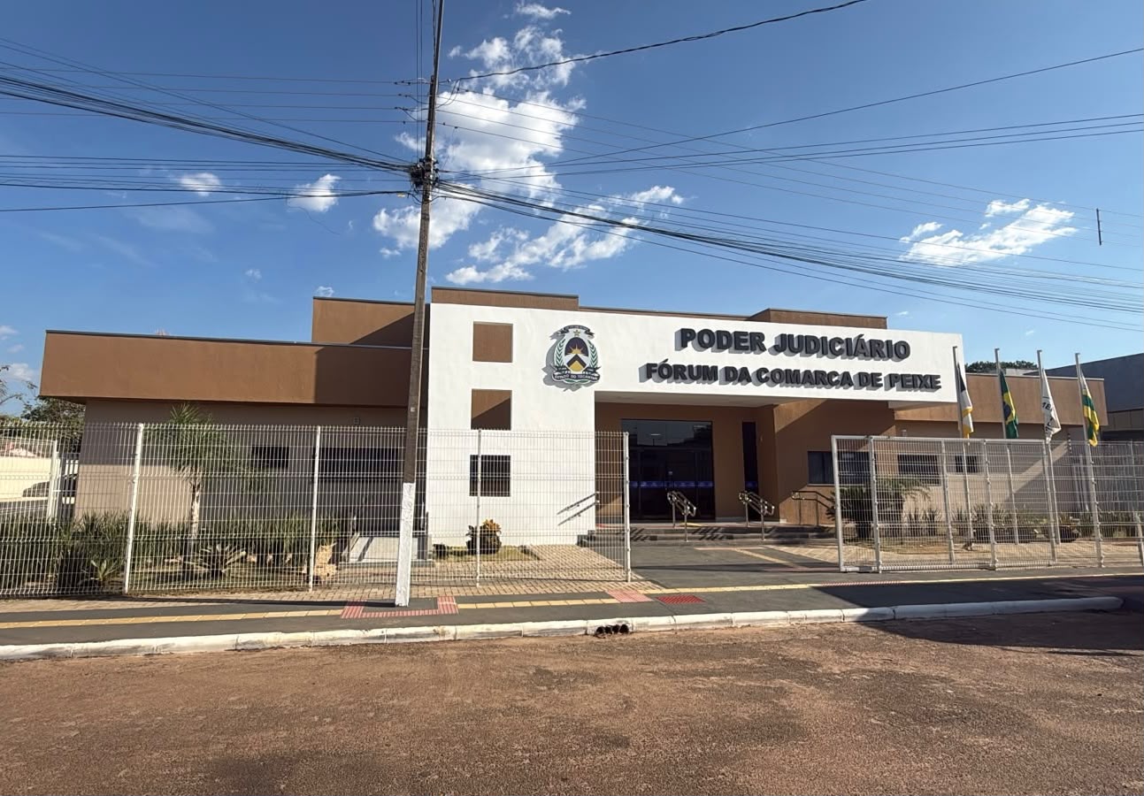 Vista frontal do Fórum da Comarca de Peixe, com fachada nas cores branca e marrom, identificação do Poder Judiciário e brasão do Estado do Tocantins. O prédio possui entrada com rampas de acesso, grades de proteção e bandeiras posicionadas na área externa.