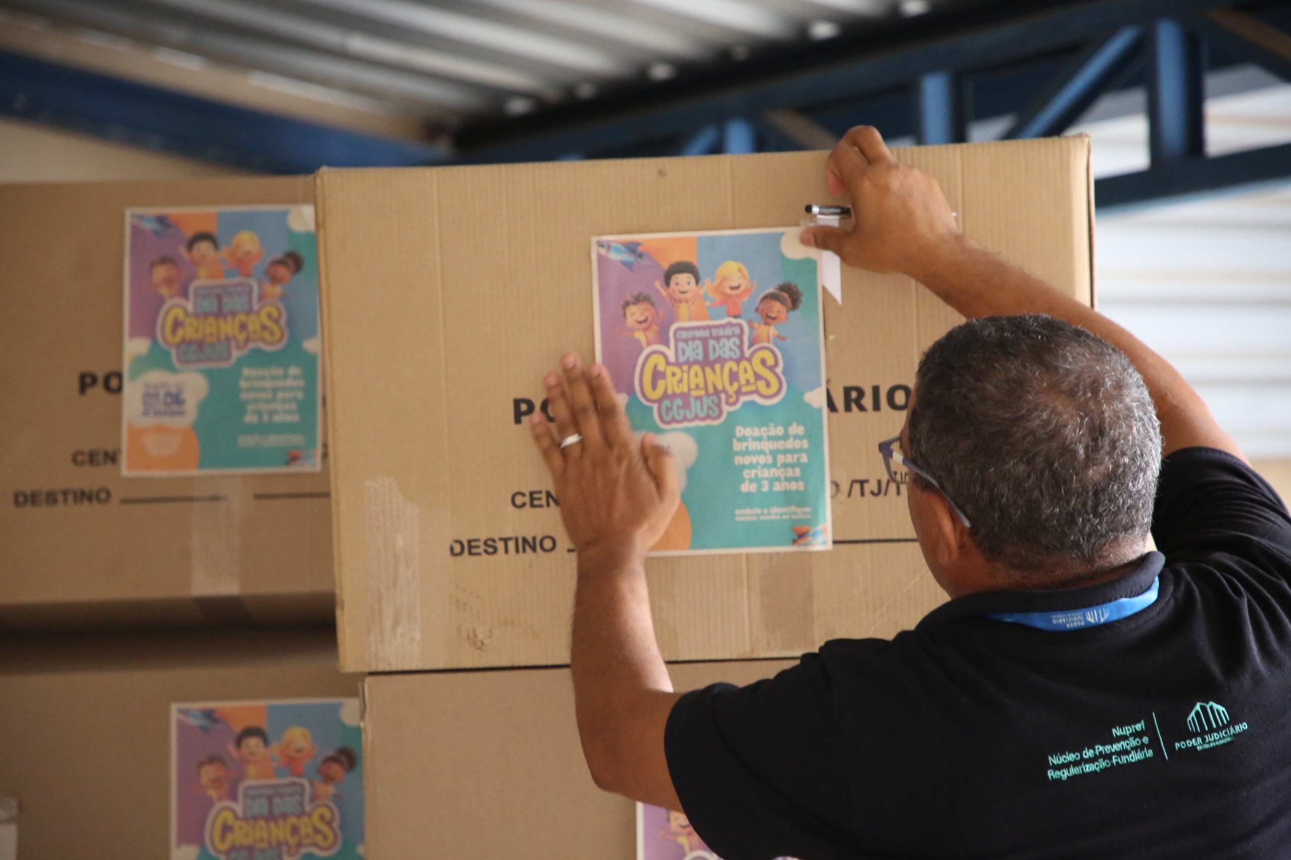 Homem de costas cola um cartaz da campanha "Dia das Crianças CGJUS" em uma caixa de papelão para arrecadação de doações.