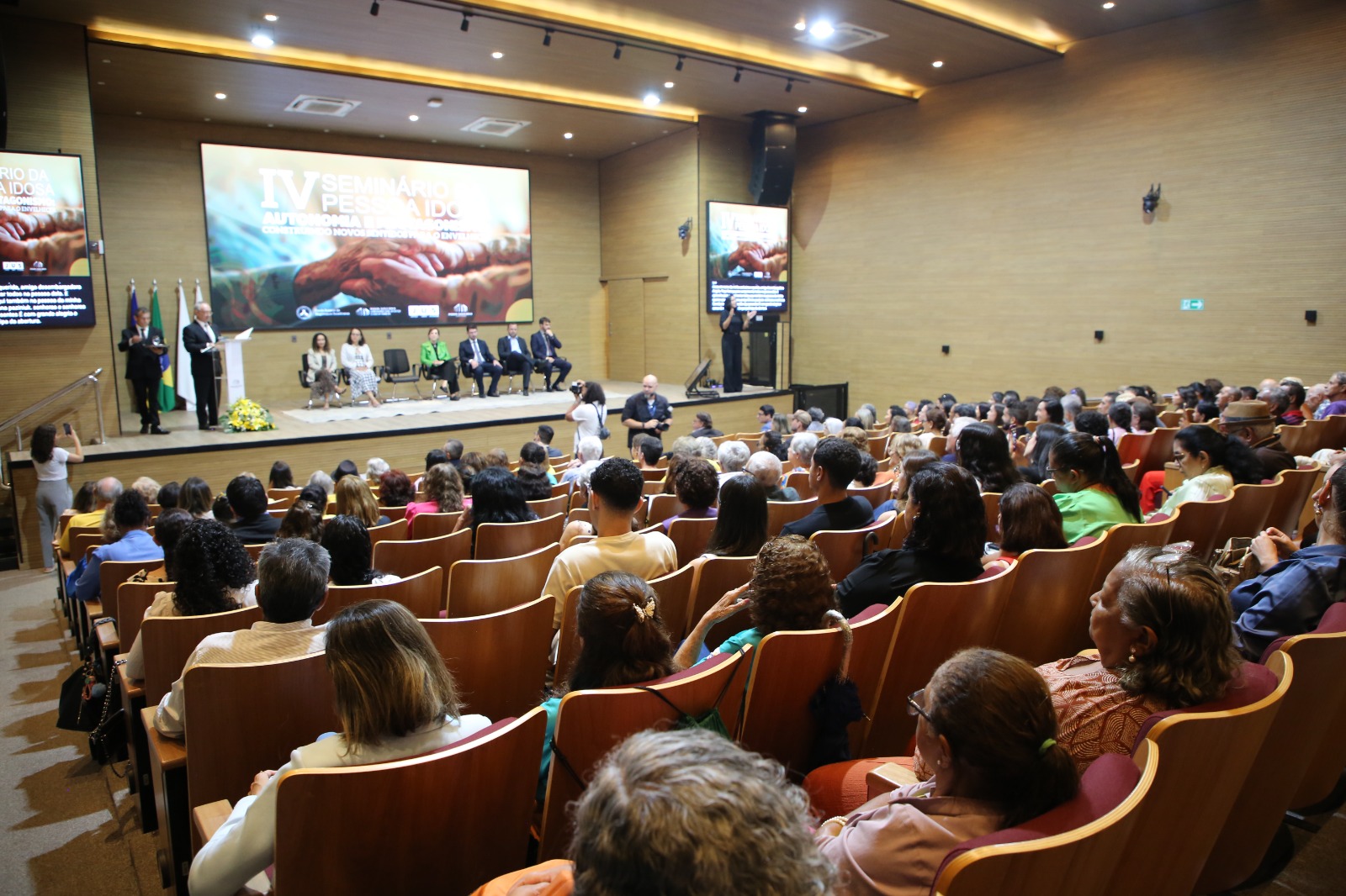 A imagem mostra o auditório do Tribunal de Justiça lotado durante a abertura do IV Seminário da Pessoa Idosa, com o público atento assistindo à apresentação no palco.