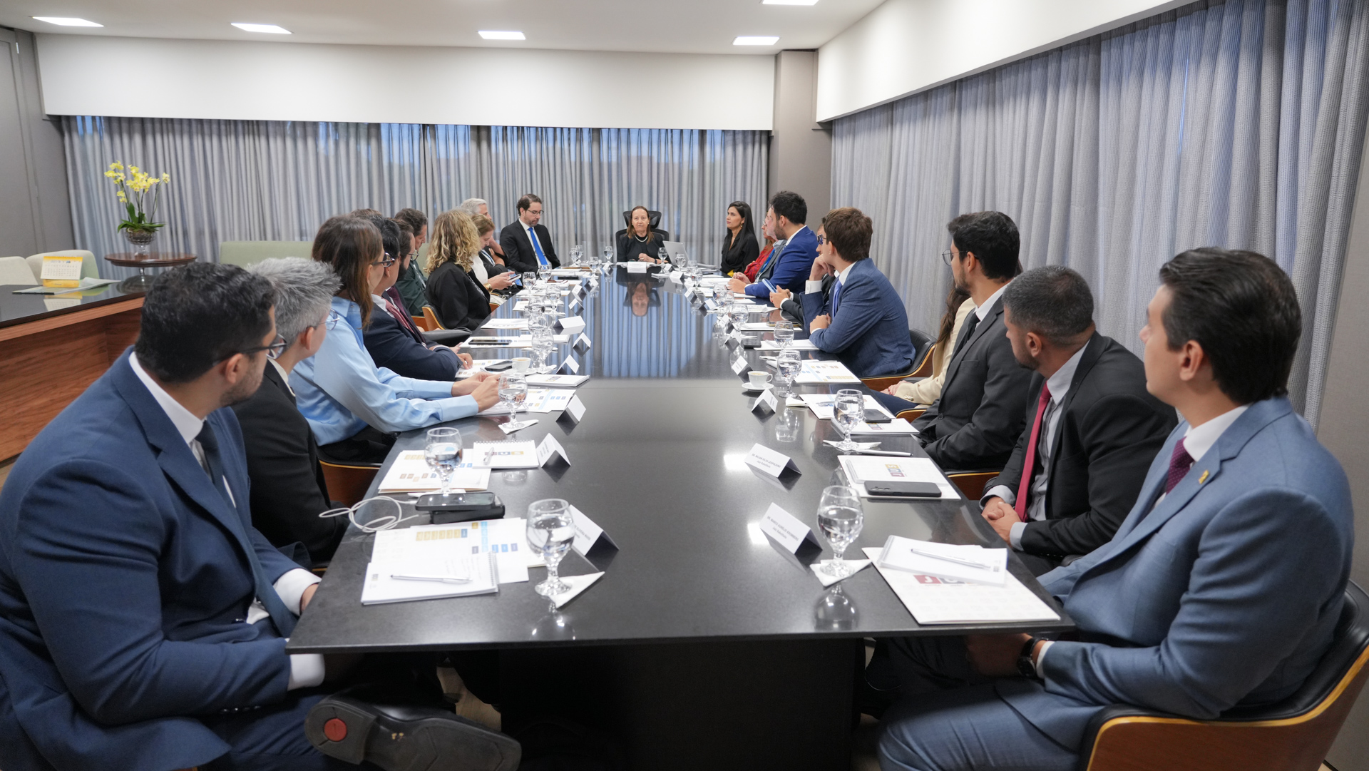 A presidente do TJTO recebe os novos juízes e juízas para apresentação da estrutura do Poder Judiciário do Tocantins. O grupo está sentado ao redor de uma longa mesa preta, com a presidente à cabeceira. Sobre a mesa há documentos, placas de identificação, celulares, taças com água e xícaras. Ao fundo, cortinas cinza cobrem as janelas.
