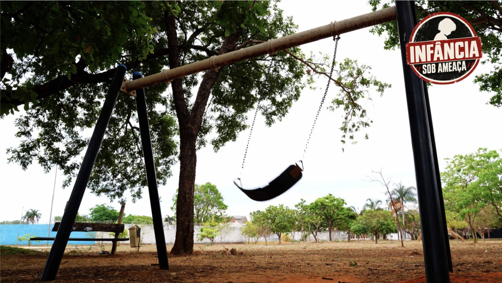 Balanço vazio, com assento preto suspenso por correntes, em uma área arborizada. Ao fundo, há um banco e um muro, sobre chão de terra coberto por folhas secas.