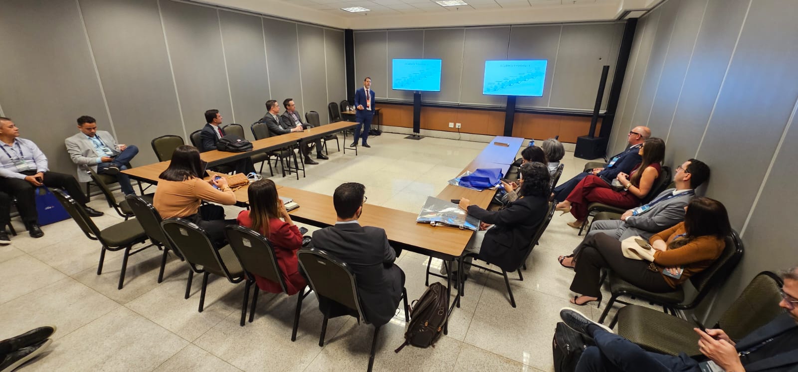 Angelo Stacciarini Seraphin, de terno azul, apresenta projetos do TJTO durante o Expojud, em Brasília. Ele está em pé, ao lado de duas telas, diante de participantes sentados ao redor de mesas dispostas em formato de U.