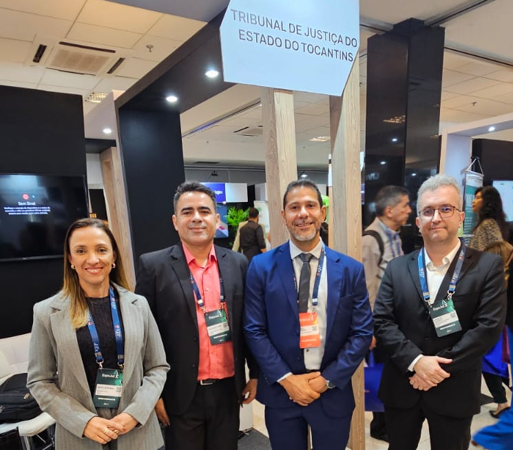 Equipe de Tecnologia da Informação do TJTO posa no estande do Tribunal durante o Expojud. Uma mulher e três homens, com roupas sociais e crachás, estão lado a lado. Acima deles, uma placa identifica o Tribunal de Justiça do Tocantins.
