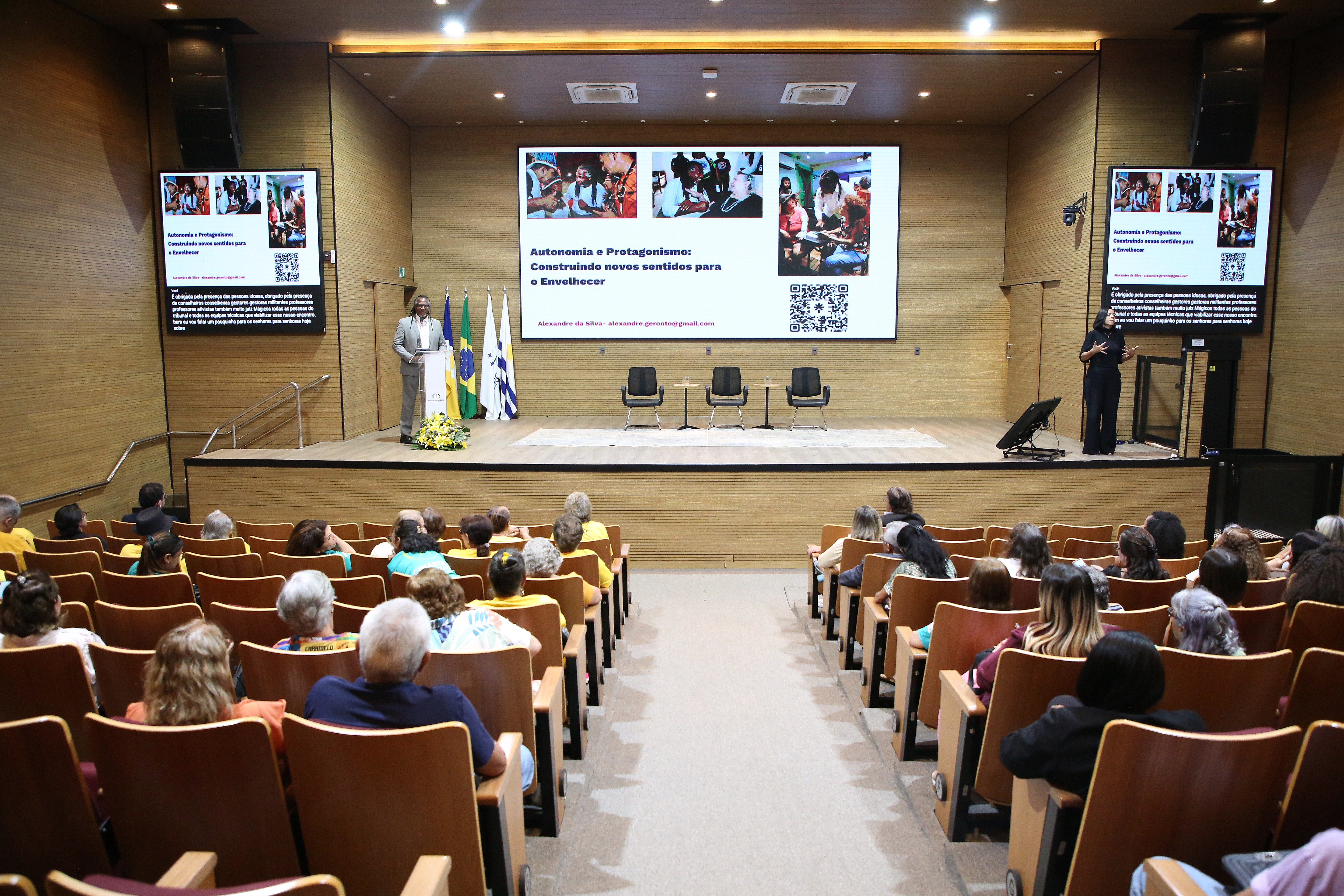 A imagem mostra a vista geral do auditório lotado durante o IV Seminário da Pessoa Idosa, com a plateia acompanhando a palestra. No palco, o palestrante discursa no púlpito ao lado de bandeiras, com um grande telão exibindo a apresentação sobre "Autonomia e Protagonismo".
