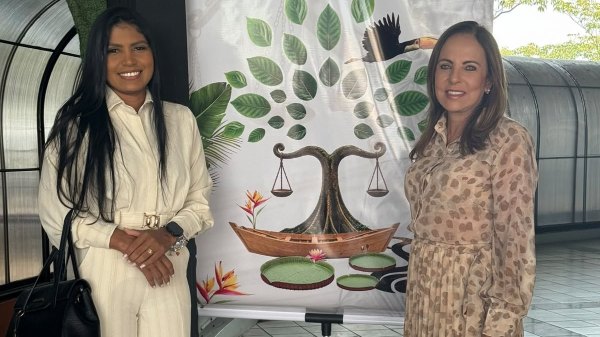 A engenheira ambiental Cinthia Barbosa (à esquerda), usando conjunto bege de manga longa, e a desembargadora Ângela Prudente (à direita), de vestido estampado, sorriem lado a lado. Entre elas há um banner da sustentabilidade, que exibe a balança da justiça integrada à natureza.