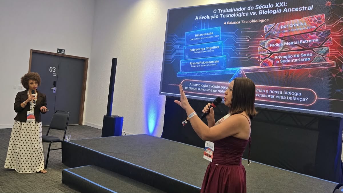 A imagem mostra duas mulheres durante uma apresentação em um evento. Uma delas segura um microfone e gesticula diante de uma tela com informações sobre tecnologia, saúde mental e riscos do excesso de conectividade no trabalho.