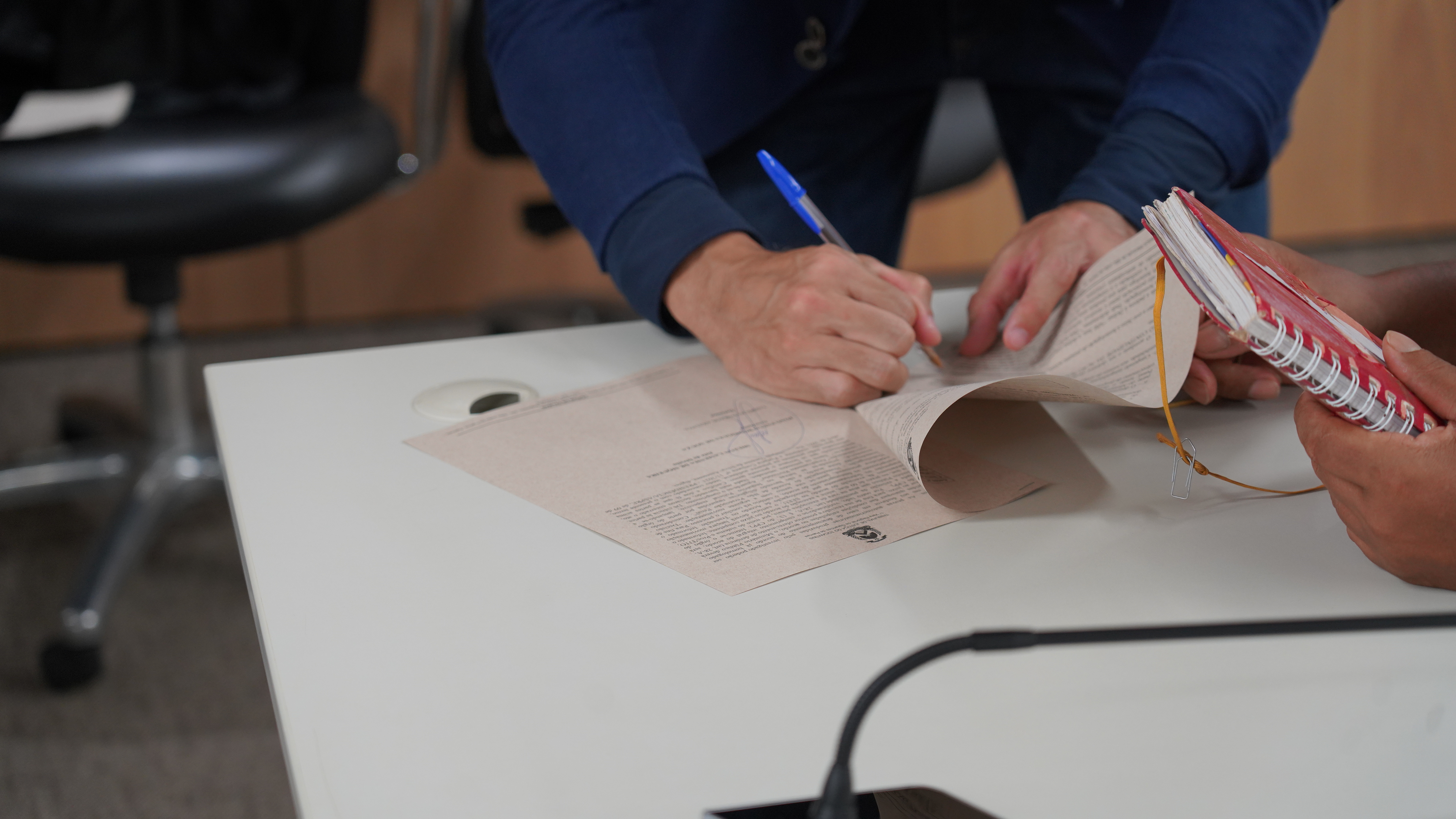 A imagem mostra uma pessoa assinando um documento impresso sobre uma mesa, enquanto outra segura um caderno. O ato foi durante a segunda edição do mutirão de ANPP.