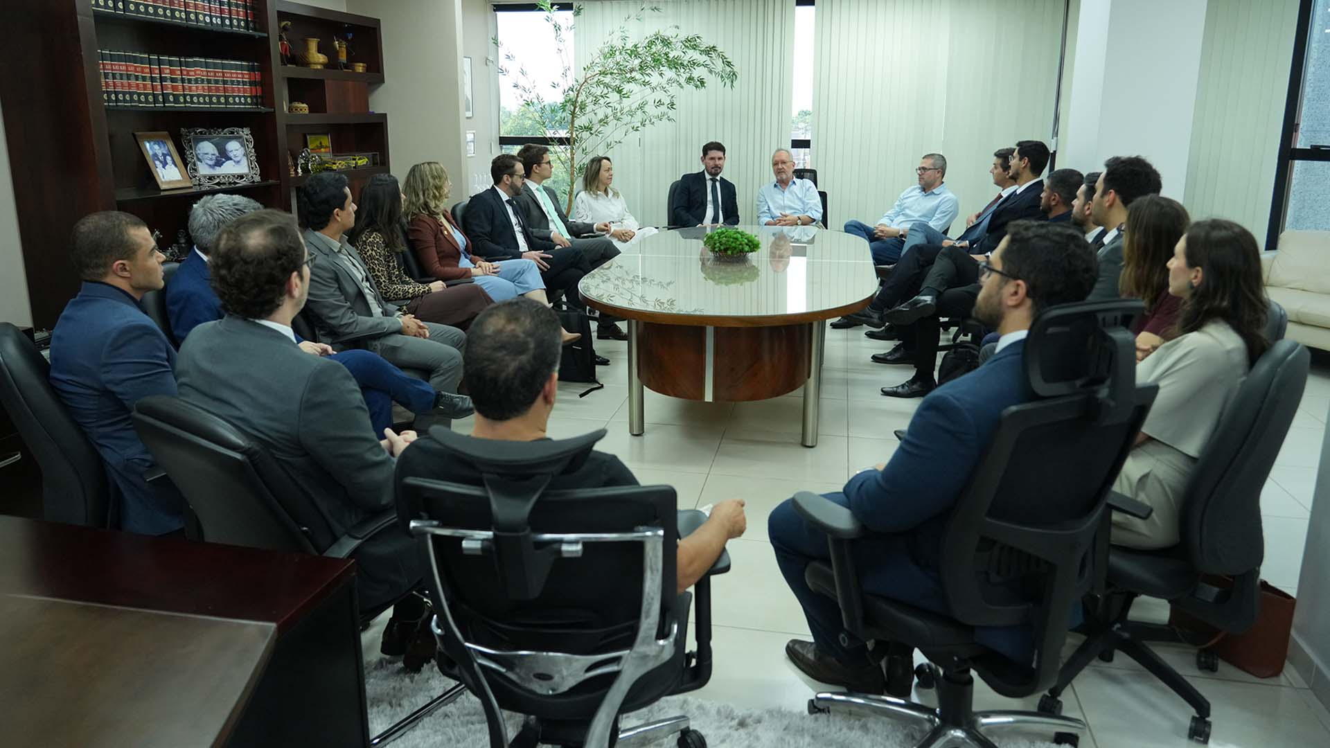 A imagem mostra um grupo de pessoas de traje formal e social reunidas em uma sala de reuniões iluminada. Elas estão dispostas em círculo ao redor de uma grande mesa oval de madeira.