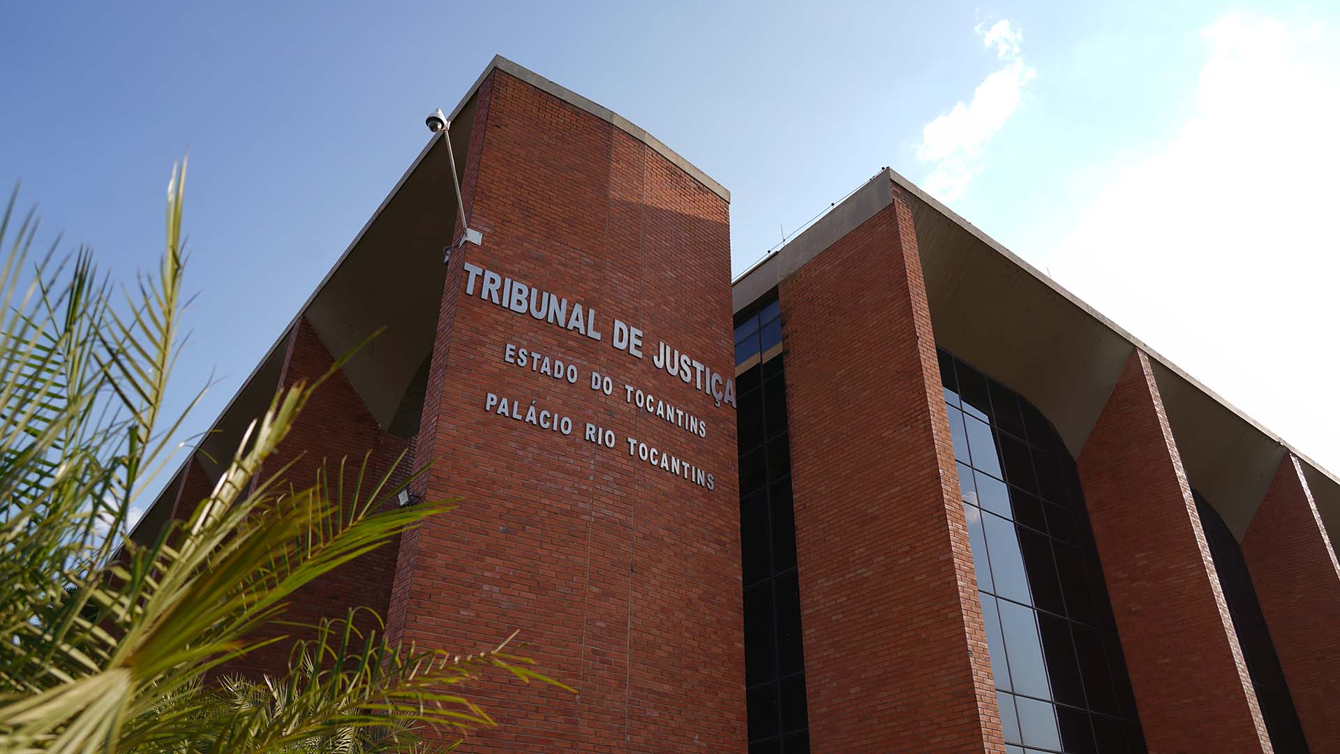 Fachada moderna do prédio do Tribunal de Justiça do Estado do Tocantins - Palácio Rio Tocantins, revestida em tijolos à vista vermelhos e grandes janelas espelhadas escuras. A foto está em ângulo inferior sob céu limpo e ensolarado, com detalhes de vegetação de palmeira em primeiro plano à esquerda.