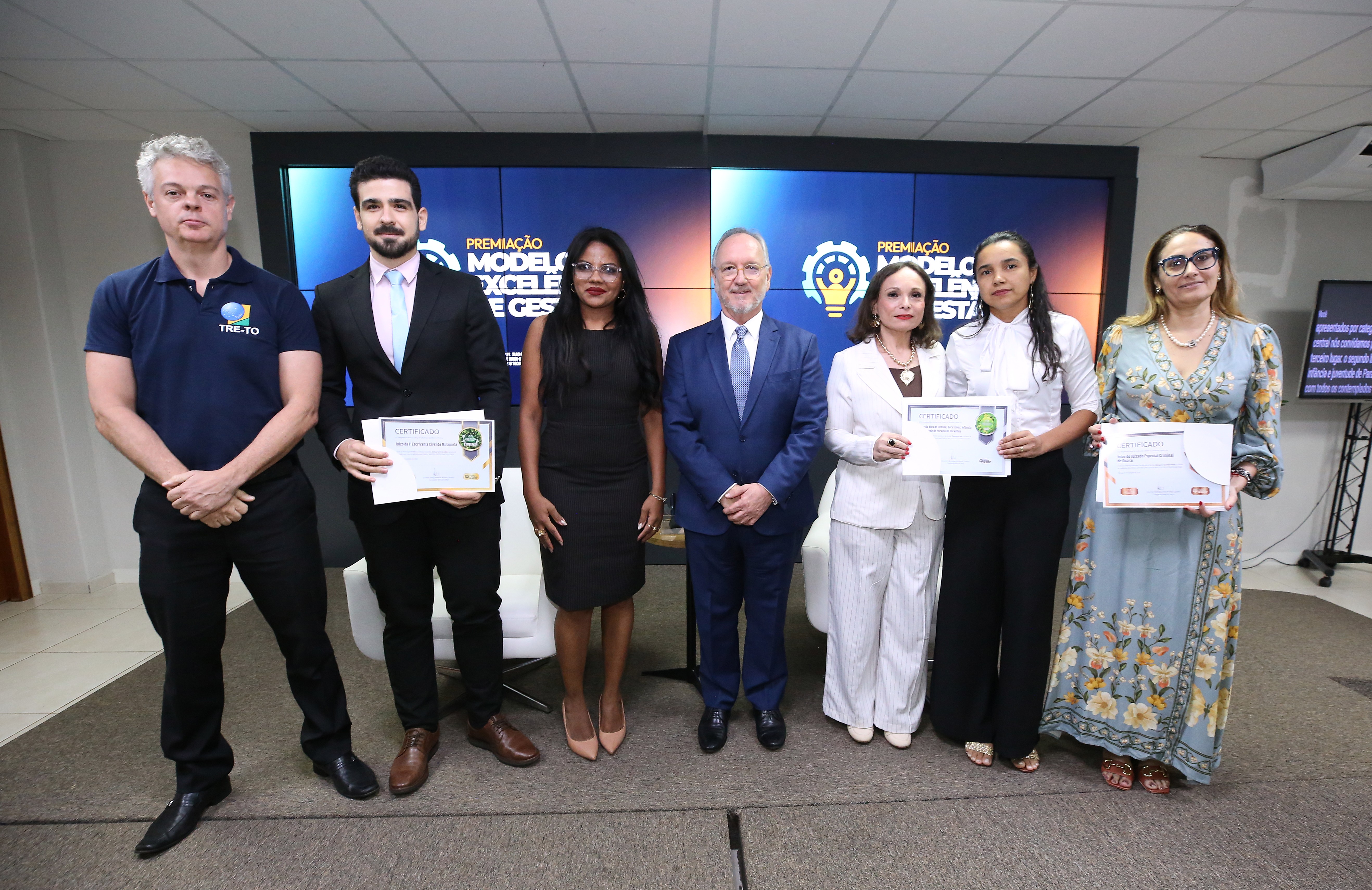 Fotografia de sete pessoas lado a lado em um auditório, três delas segurando certificados de premiação. Ao fundo, telas exibem a marca do evento "Premiação Modelo de Excelência em Gestão".