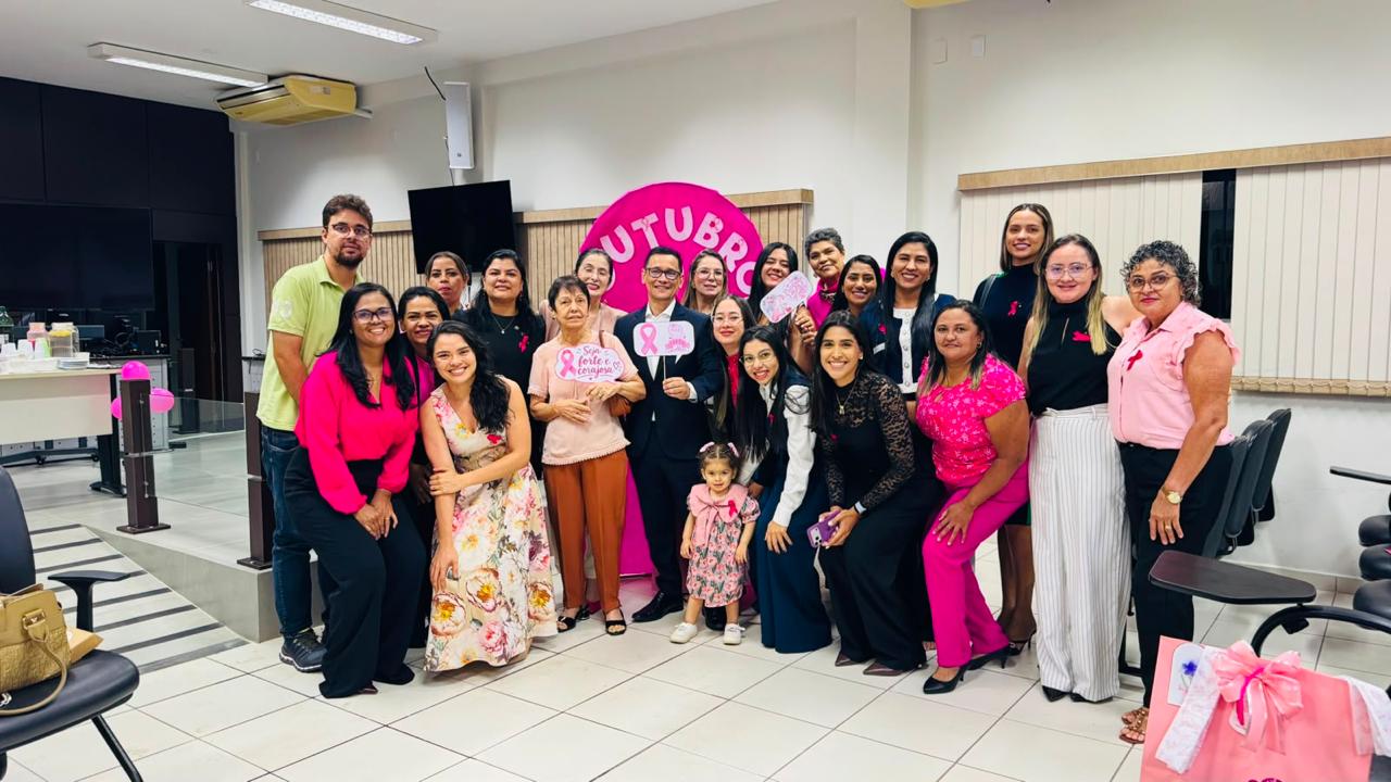 A imagem mostra um grupo de mulheres e homens reunidos no Fórum da Comarca de Augustinópolis para uma ação do Outubro Rosa. Ao centro, os participantes seguram cartazes com mensagens de conscientização sobre o câncer de mama. Ao fundo, há um painel rosa alusivo à campanha. Várias pessoas vestem roupas nas cores rosa e preto, em referência à iniciativa de prevenção e valorização da saúde da mulher.