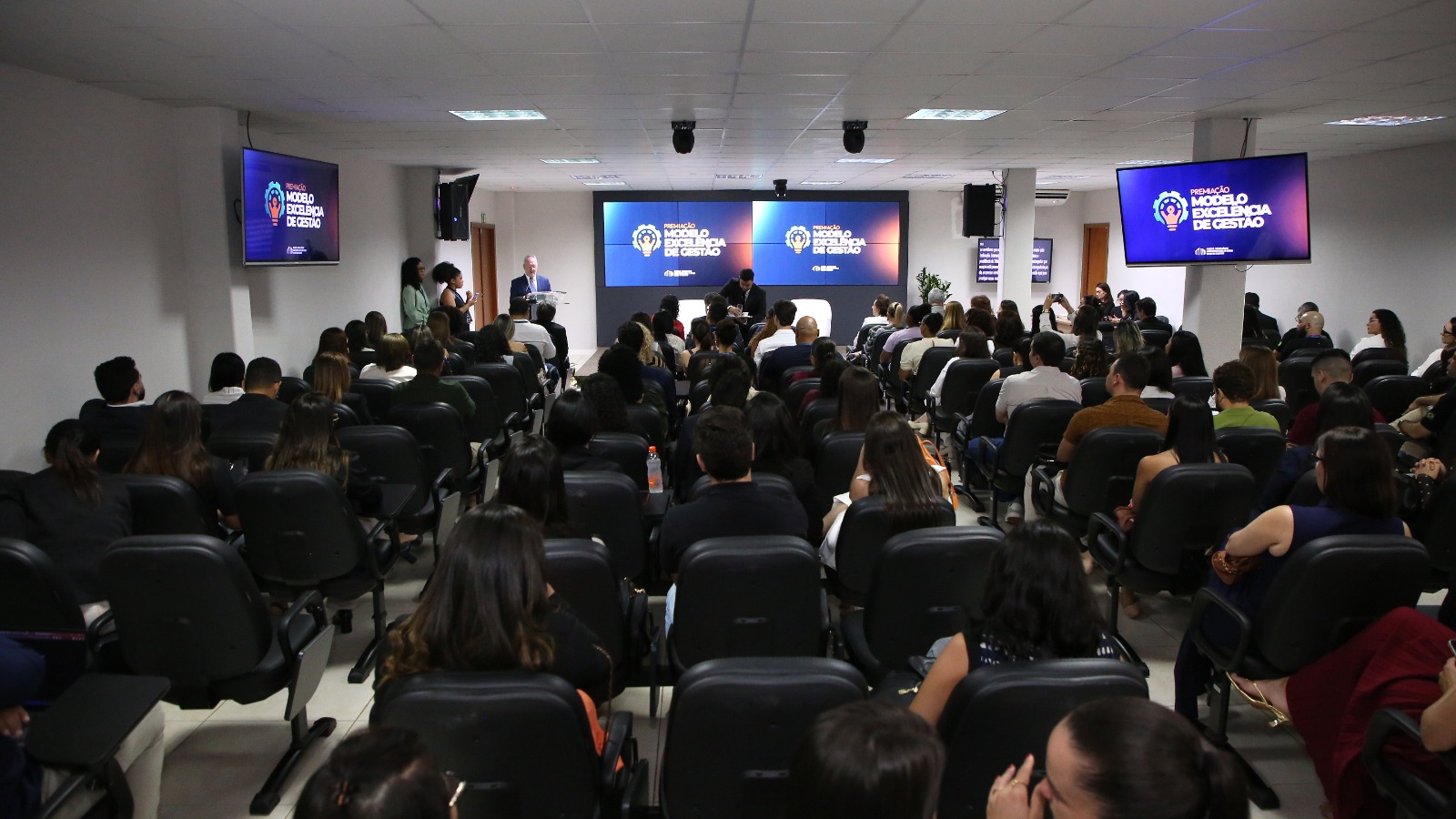 A imagem mostra um auditório com várias pessoas sentadas, acompanhando uma apresentação sobre o Prêmio Modelo de Excelência de Gestão, exibido nas telas ao fundo e nas laterais. À frente, um homem fala ao público em um púlpito.