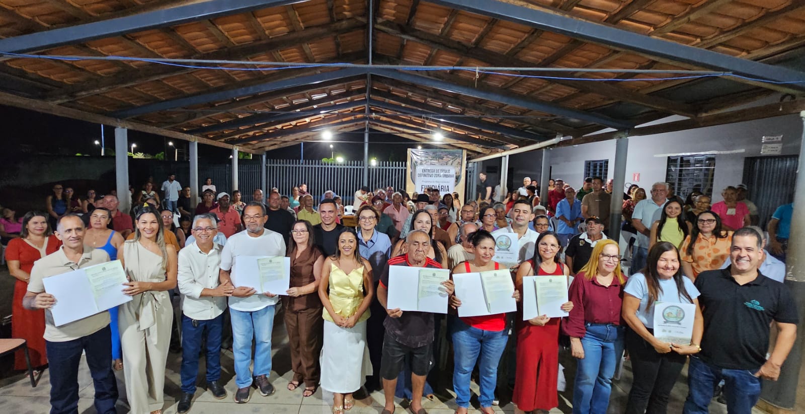 Imagem mostra várias pessoas em pé segurando títulos de propriedade sob um barracão