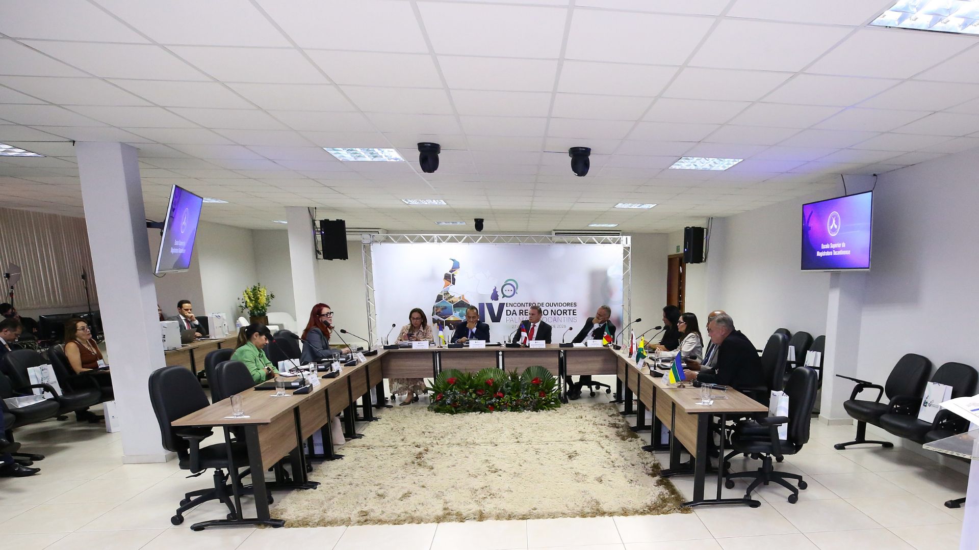 Foto mostra participantes do IV Encontro de Ouvidores da Região Norte reunidos em uma sala, sentados ao redor de mesas dispostas em formato de U. Ao centro, autoridades compõem a mesa principal. Ao fundo, um painel identifica o evento, e arranjos de flores decoram o espaço. Bandeiras dos estados da Região Norte estão posicionadas sobre as mesas.