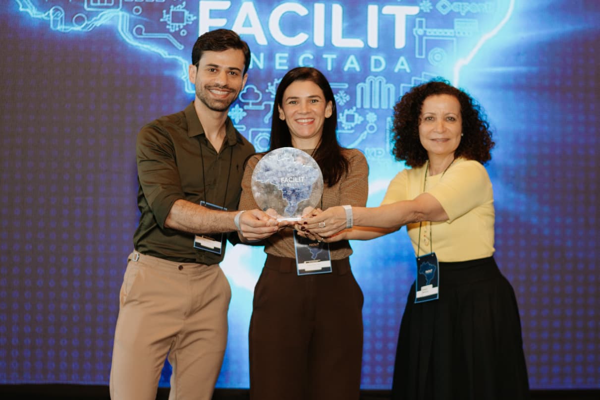 Três participantes posam para foto segurando um troféu durante evento institucional. Ao fundo, um painel iluminado com a inscrição “Facilit Conectada” remete ao tema de tecnologia e inovação