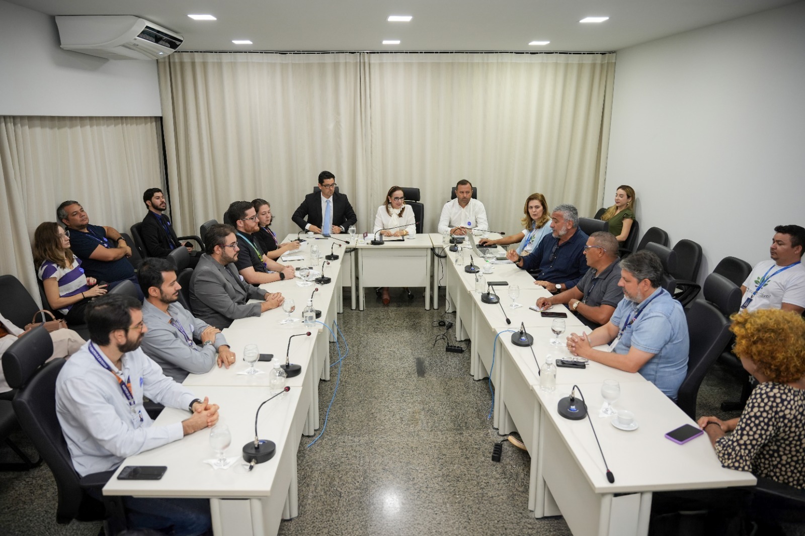 Homens e mulheres sentados em mesa branca em formato U, em sala de paredes brancas e persiana bege