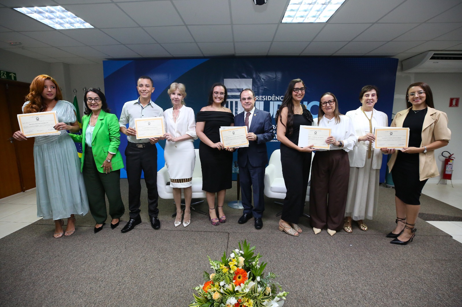A imagem mostra um grupo de pessoas posando para uma foto na Esmat. Elas seguram os certificados da Turma III do Programa de Residência em Prática Jurídica. Ao fundo, há um painel oficial e, à frente, um arranjo de flores, compondo um cenário formal de solenidade.