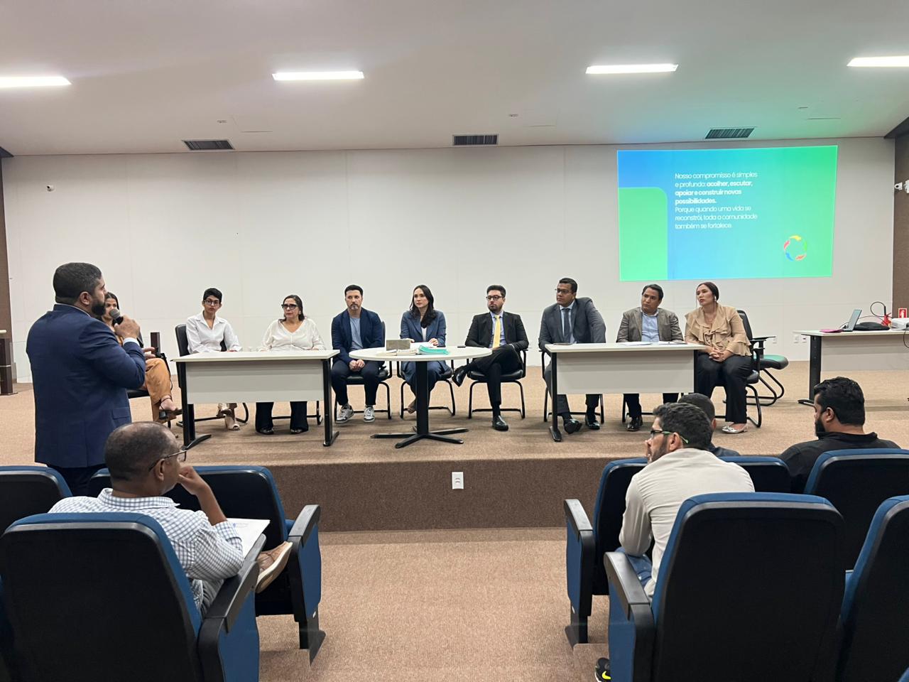 autoridades participam de evento. Todos estão sentados e mesas brancas aparecem na frente