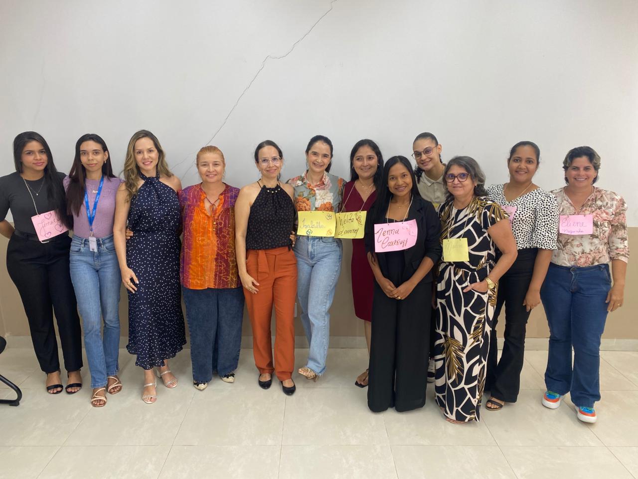 Mulheres estão em pé, alinhadas lado a lado, posando para a foto diante de uma parede branca. Algumas usam papéis coloridos com nomes escritos à mão presos à roupa. Todas olham para a câmera e sorriem. O ambiente é interno, com piso claro e sem elementos decorativos ao fundo.