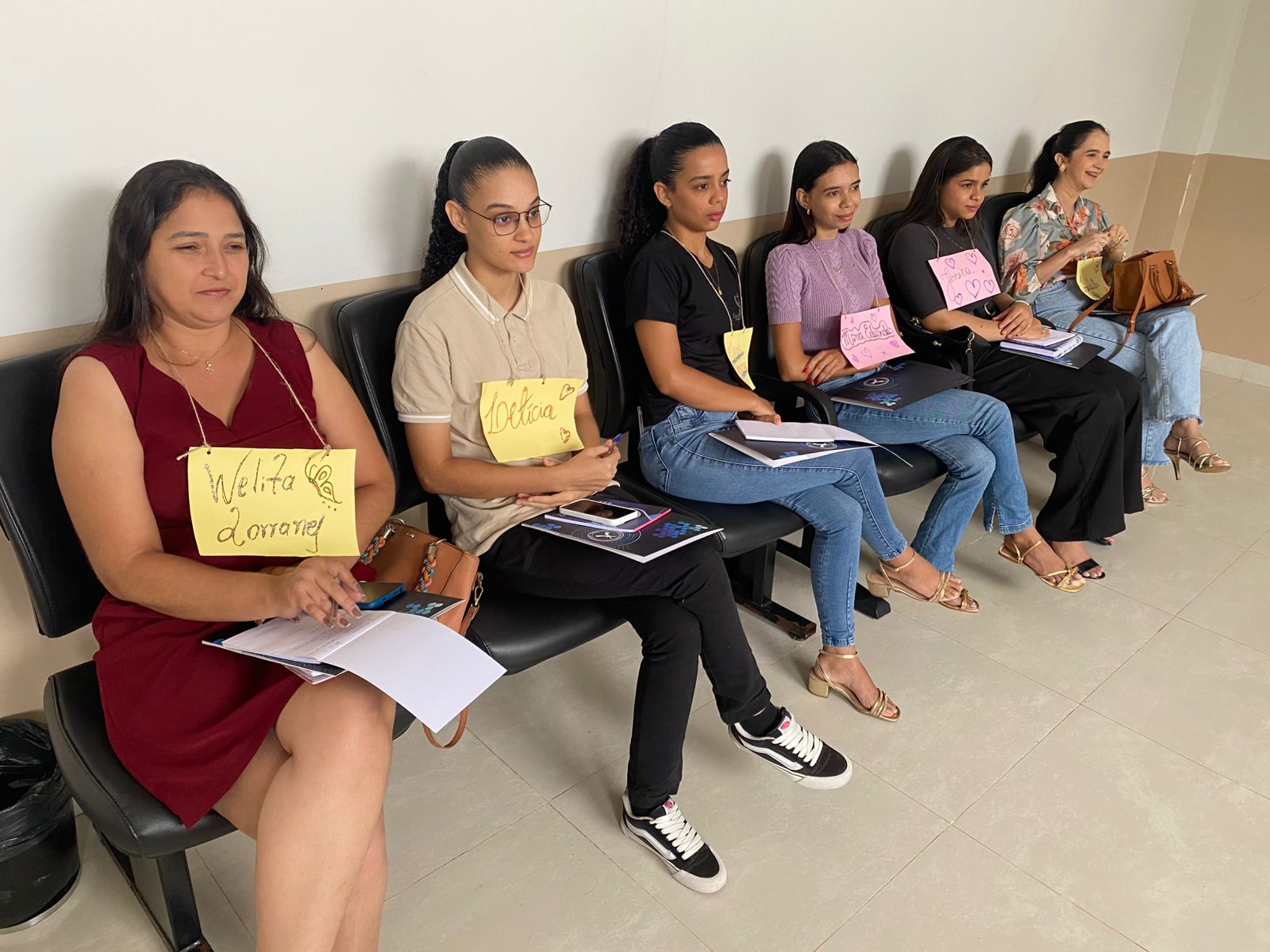 Seis mulheres estão sentadas lado a lado em cadeiras pretas encostadas na parede, em ambiente interno. Todas usam papéis coloridos com nomes escritos à mão pendurados no pescoço.  Algumas seguram cadernos, folhas e canetas sobre o colo. O espaço possui parede clara e piso de cerâmica clara.