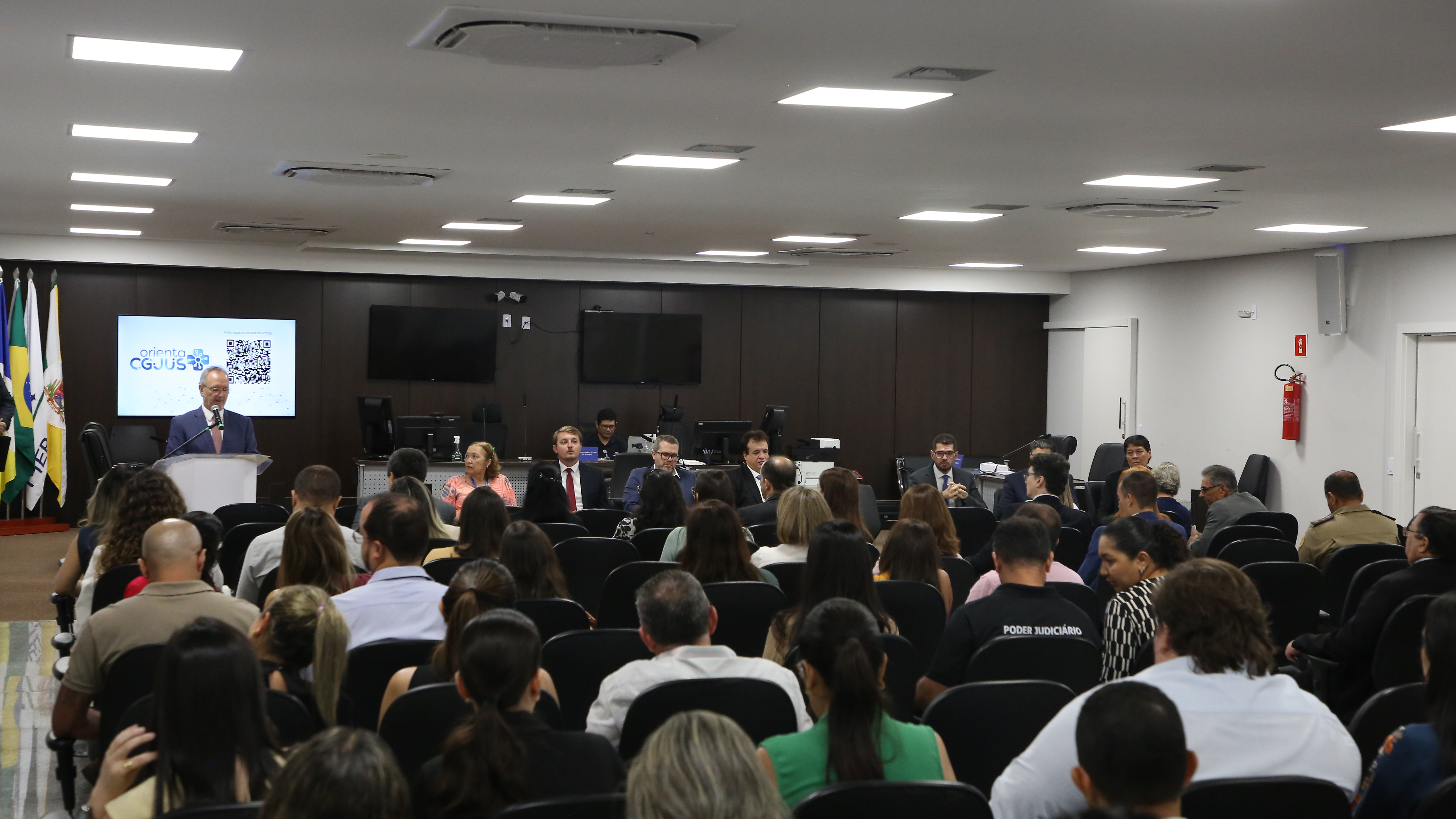 Uma sala de audiência ou auditório institucional com várias pessoas sentadas em cadeiras organizadas em fileiras, assistindo a uma apresentação. Na frente, um homem fala ao púlpito com microfone, ao lado de uma tela que exibe um slide com QR code e logomarca institucional.