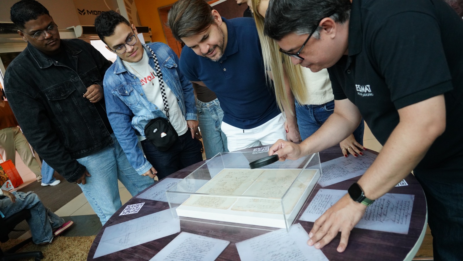 Grupo de pessoas observa atentamente documentos antigos expostos em uma mesa, protegidos por uma caixa transparente, enquanto um homem usa uma lupa para mostrar detalhes do material em uma exposição