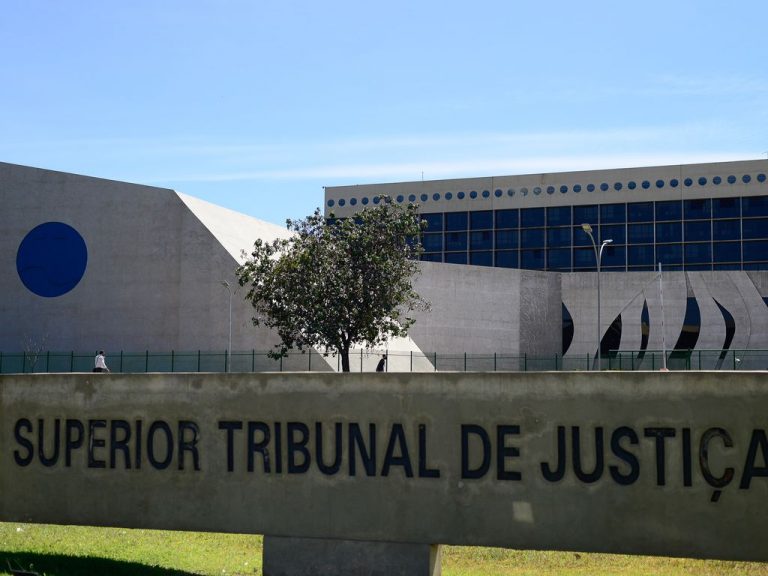 Sede do Superior Tribunal de Justiça, em Brasília.  Em primeiro plano, monumento de concreto com a inscrição do nome do tribunal. Ao fundo, prédio de arquitetura moderna, com fachada em concreto claro e painéis de vidro azul, área gramada e céu aberto.