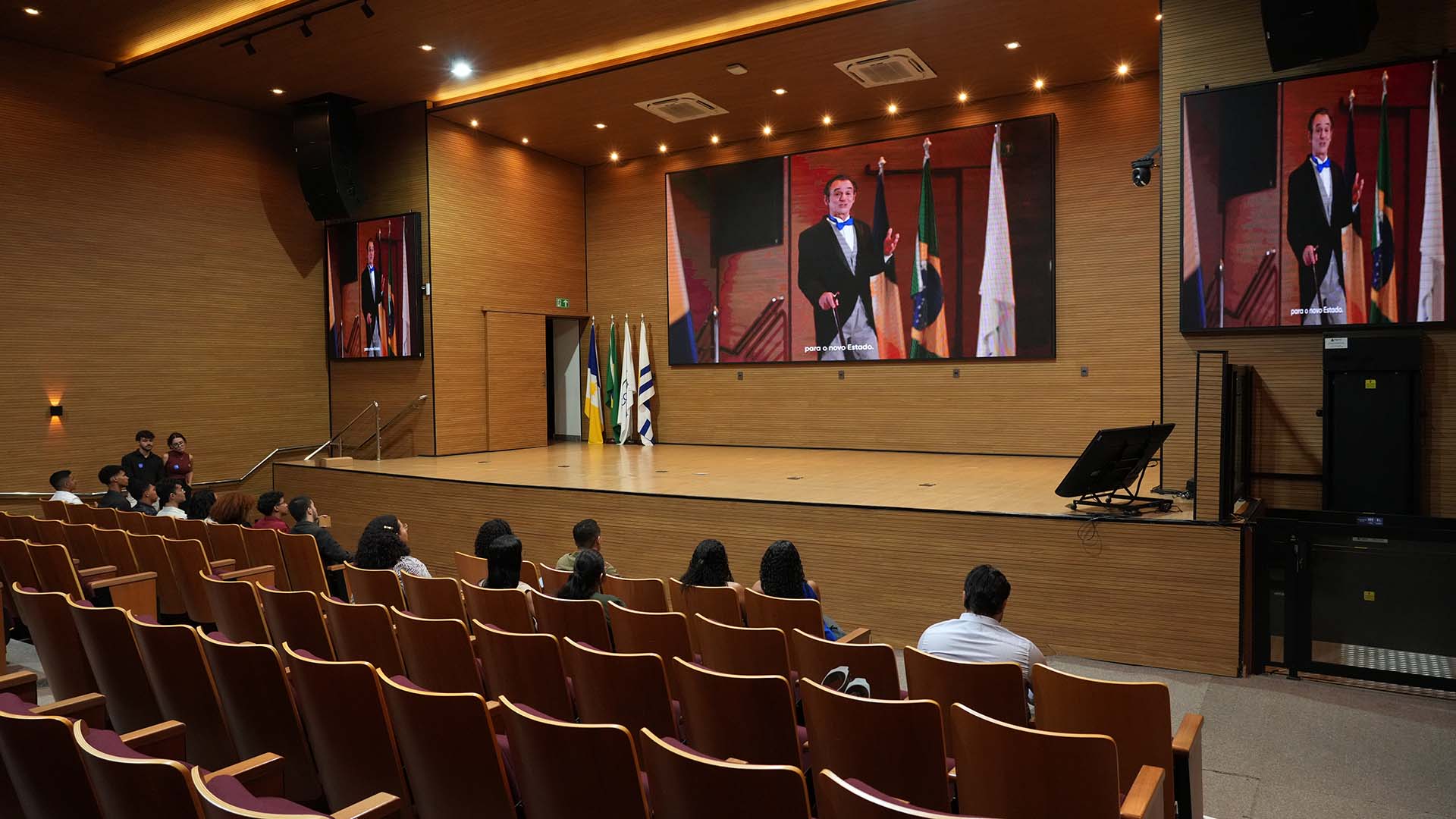 Auditório com palco e três telões exibindo um vídeo de um homem discursando diante de bandeiras. Algumas pessoas assistem sentadas na plateia. Ambiente com revestimento em madeira e estrutura audiovisual instalada.