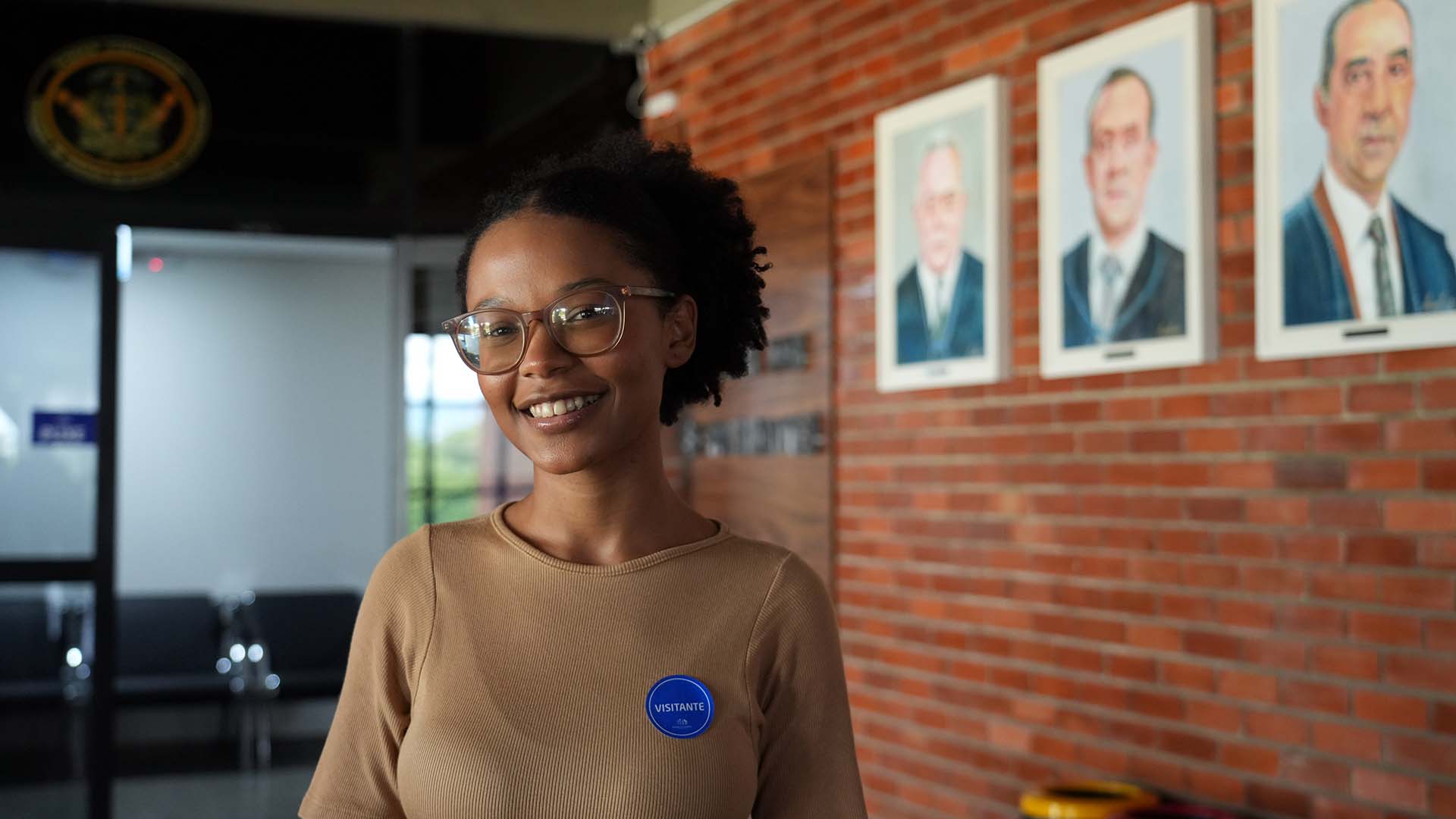 Jovem, usando óculos e roupa bege com adesivo “Visitante”, sorri para a câmera. Ao fundo, parede de tijolos com retratos emoldurados e área interna de recepção.