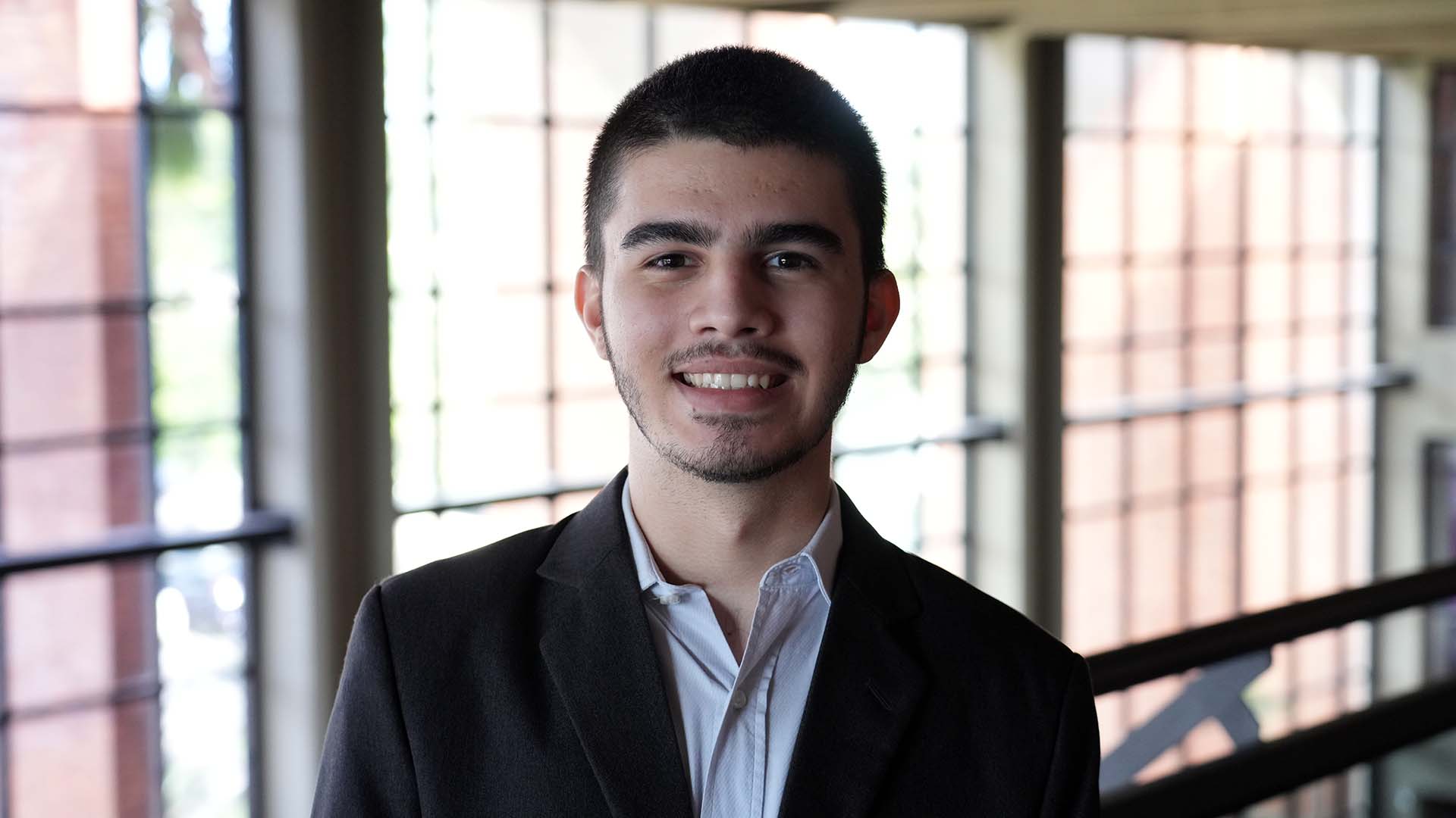 Jovem, com cabelo curto e barba discreta, veste blazer preto e camisa branca. Ele sorri para a câmera em ambiente interno do TJTO, com janelas amplas ao fundo