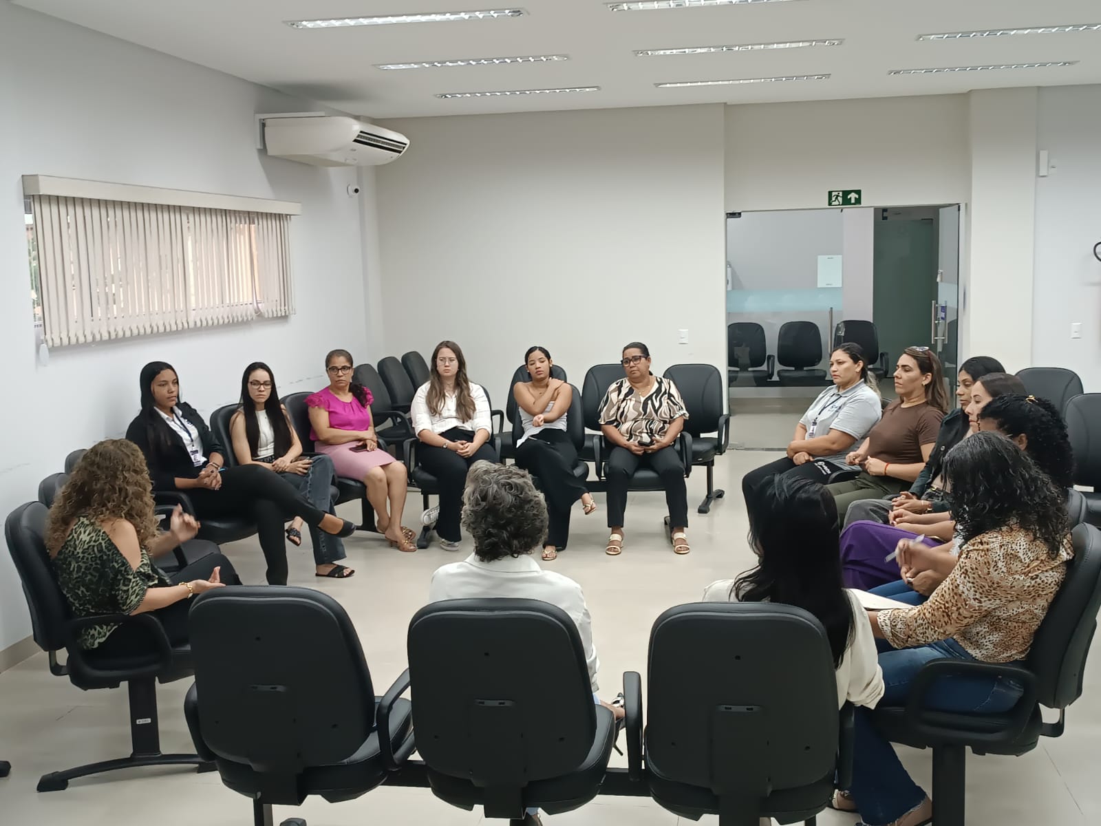 Servidoras participam de roda de conversa conduzida pela equipe do NAPsi na Comarca de Taguatinga. Sentadas em círculo, as participantes acompanham atentamente a exposição de uma das facilitadoras, em ambiente reservado, com cadeiras organizadas em formato colaborativo. O espaço conta com iluminação clara, ar-condicionado e porta de acesso ao fundo.