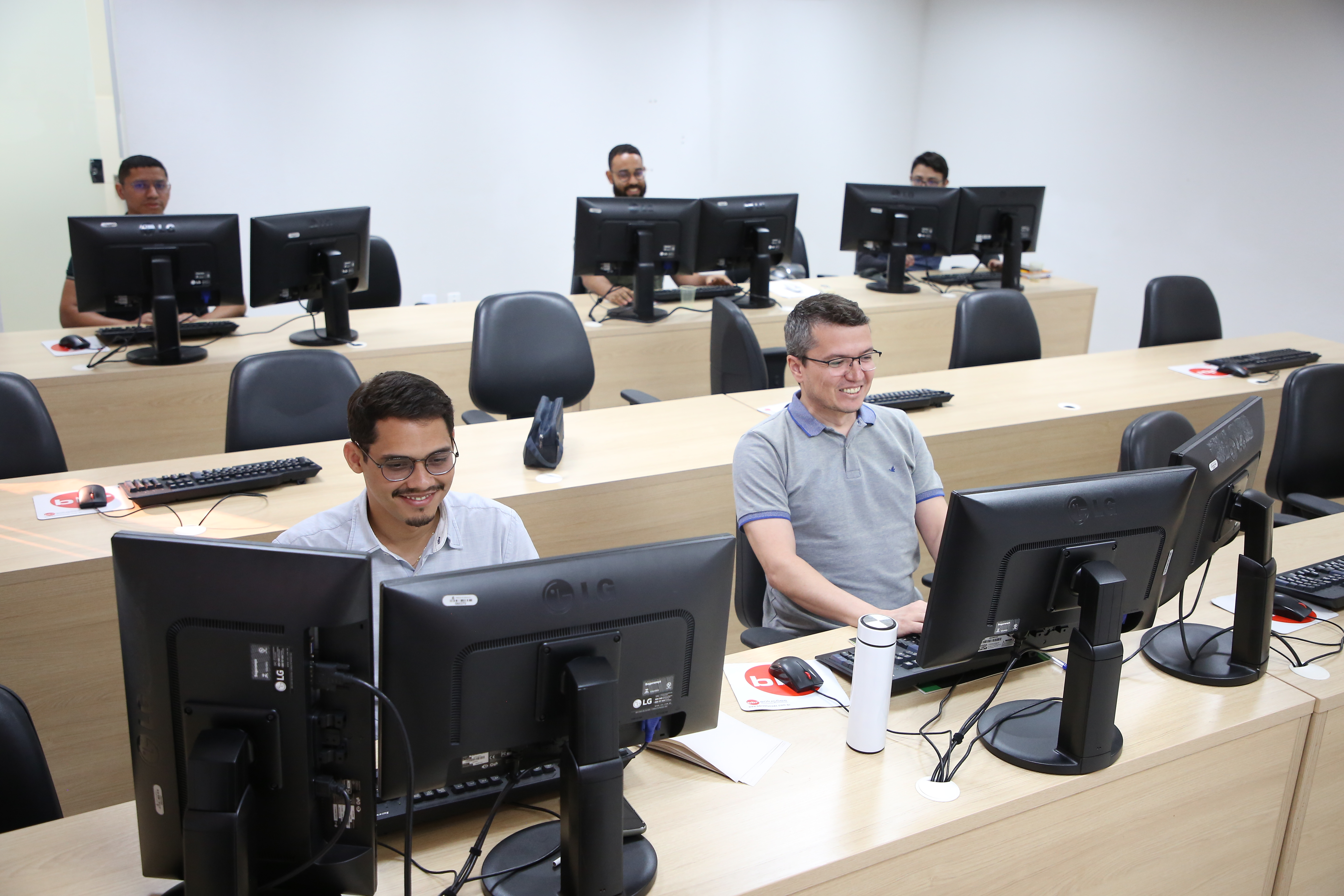 Cinco homens utilizam computadores em uma sala organizada, com mesas em fileiras e cadeiras pretas. Dois estão em destaque na primeira fileira, sorrindo diante dos monitores. O ambiente é claro, com mobiliário em tom amadeirado e estrutura de ambiente de trabalho..