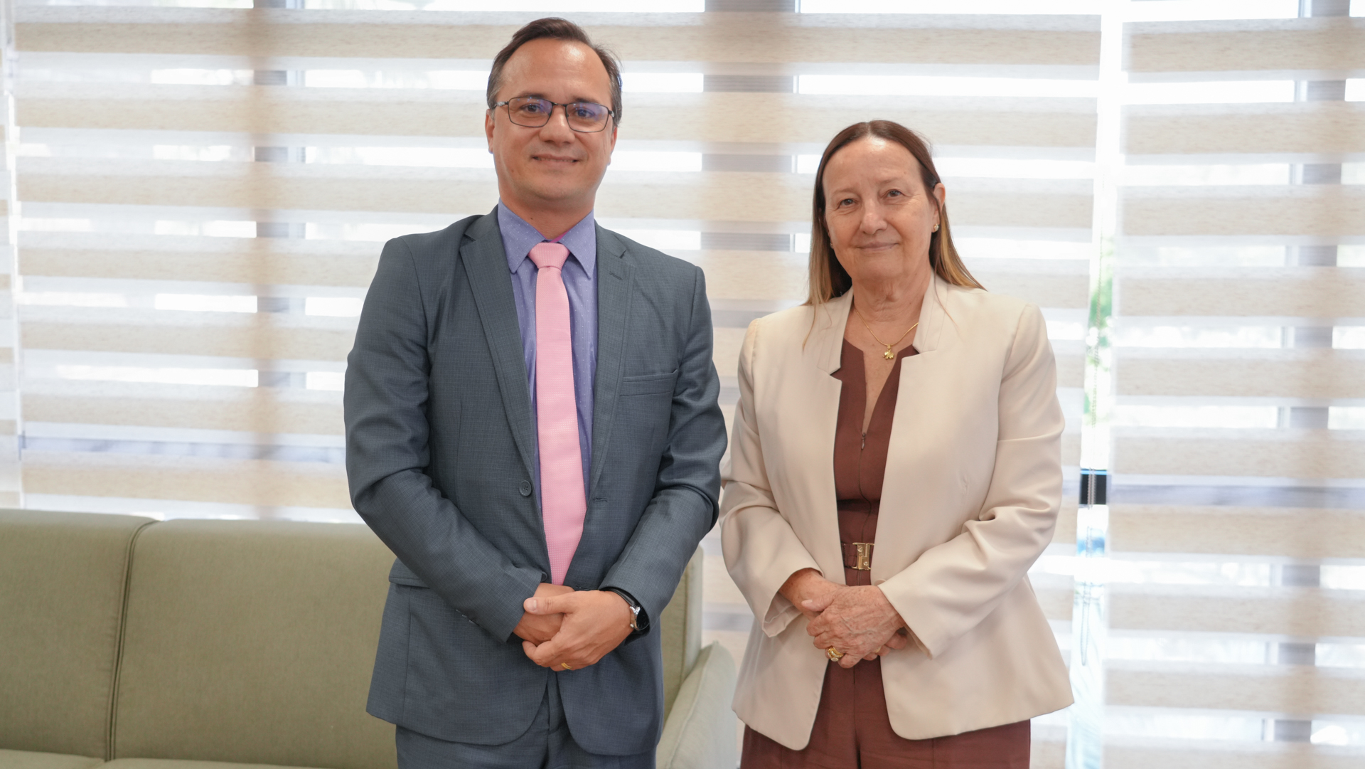 A imagem mostra duas pessoas em pé, lado a lado, em um ambiente interno com iluminação clara e fundo composto por persianas horizontais translúcidas. A cena transmite formalidade e cordialidade.  À esquerda, há um homem vestindo terno em tom cinza, camisa clara e gravata rosa. Ele usa óculos e mantém as mãos unidas à frente do corpo, com postura ereta e expressão serena, olhando diretamente para a câmera.  À direita, uma mulher usa blazer claro sobre vestido em tom marrom. Ela também mantém as mãos unidas à frente do corpo, com postura semelhante, expressão tranquila e discreto sorriso.