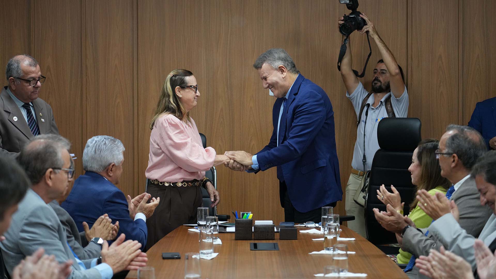 A imagem registra um momento solene em ambiente institucional. Ao centro, duas autoridades estão em pé, cumprimentando-se com um aperto de mãos sobre a mesa de reuniões. De um lado, uma mulher veste blusa em tom claro e calça escura; do outro, um homem traja terno azul, ambos com expressão cordial e respeitosa, indicando formalização de um ato ou acordo.  Ao redor da mesa, diversas pessoas estão sentadas, a maioria de trajes formais, aplaudindo o gesto central, o que reforça o caráter oficial e comemorativo da ocasião. Sobre a mesa, observam-se copos com água, papéis e materiais de escritório, típicos de uma reunião administrativa.  Ao fundo, há um fotógrafo em pé, registrando o momento, o que evidencia tratar-se de um evento oficial ou institucional. O ambiente é uma sala de reuniões com paredes revestidas em madeira, transmitindo sobriedade e formalidade compatíveis com cerimônias do Poder Público.