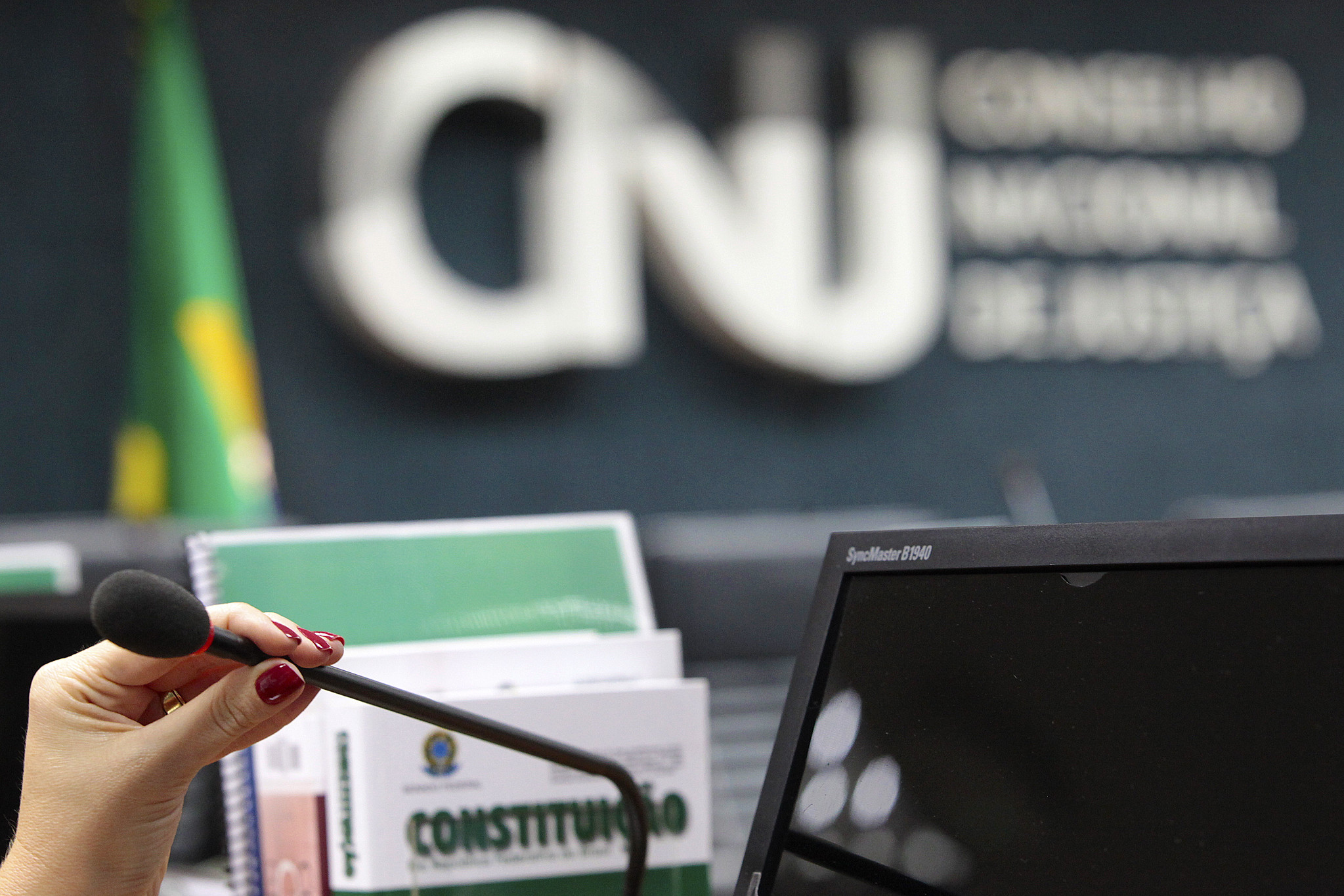 Close de uma mão feminina segurando um microfone, posicionada diante de um monitor de computador e de exemplares impressos da Constituição Federal. Ao fundo, de forma desfocada, aparece a logomarca do Conselho Nacional de Justiça (CNJ) e parte da bandeira do Brasil.
