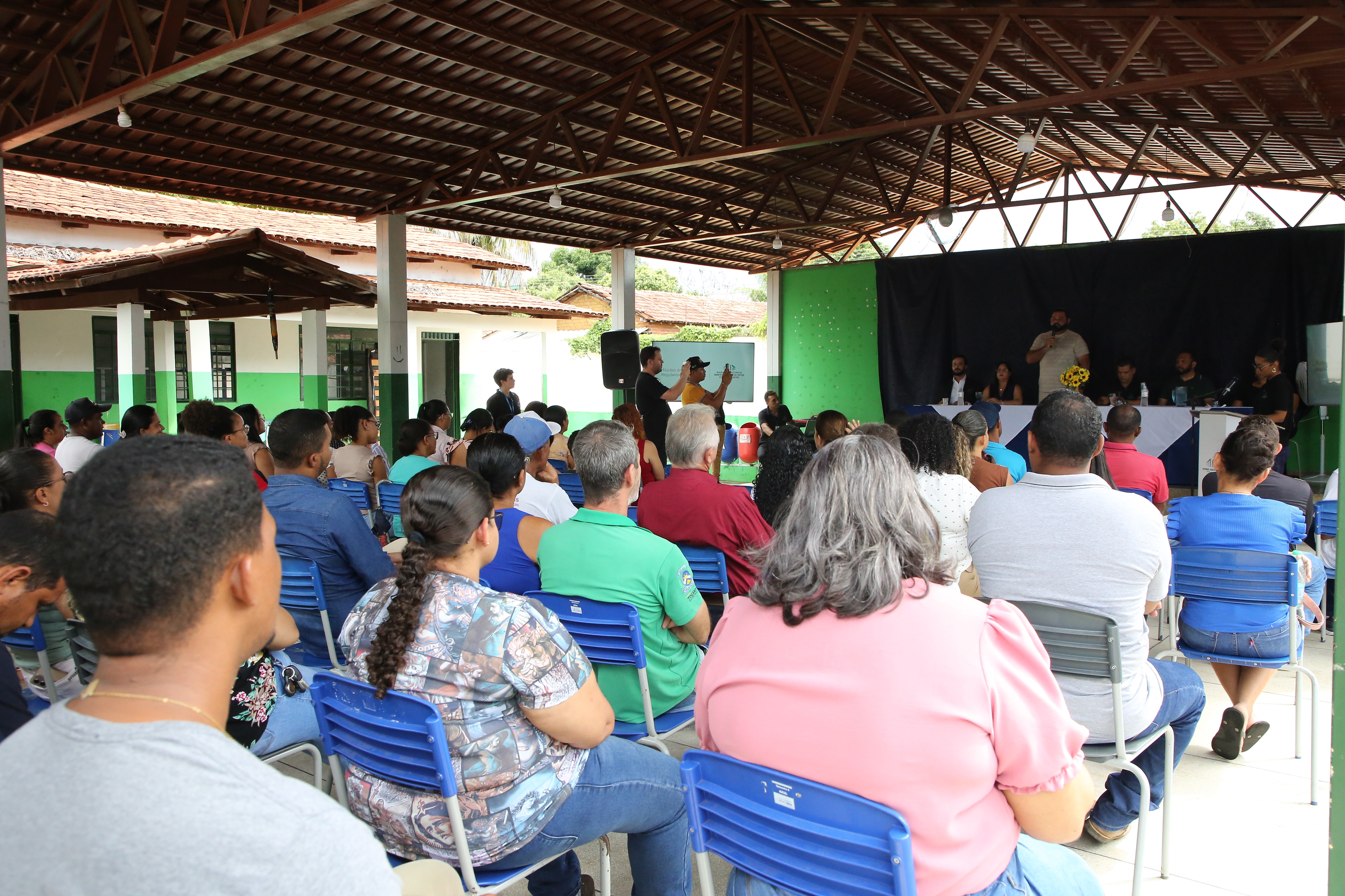 A imagem mostra uma audiência pública sobre a regularização fundiária realizada pela Corregedoria-Geral da Justiça do Tocantins, com a participação da comunidade local. Várias pessoas estão reunidas em um espaço coberto, sentadas em cadeiras azuis, acompanhando as discussões conduzidas pelas autoridades.