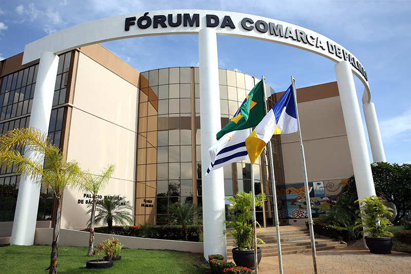 A imagem mostra a fachada do Fórum da Comarca de Palmas, edifício de arquitetura moderna e institucional. Em destaque, vê-se um grande arco branco com a inscrição “FÓRUM DA COMARCA DE PALMAS”, sustentado por pilares, marcando a entrada principal do prédio.  A construção possui linhas curvas e volumetria ampla, com fachada em tons claros, combinando superfícies lisas e áreas envidraçadas, que permitem a entrada de luz natural. À frente do edifício, há um jardim bem cuidado, com gramado, palmeiras e vasos ornamentais, reforçando o aspecto organizado e solene do espaço público.  No centro da área frontal, três mastros exibem bandeiras oficiais — do Brasil, do Estado do Tocantins e do Município — posicionadas próximas à escadaria de acesso principal.