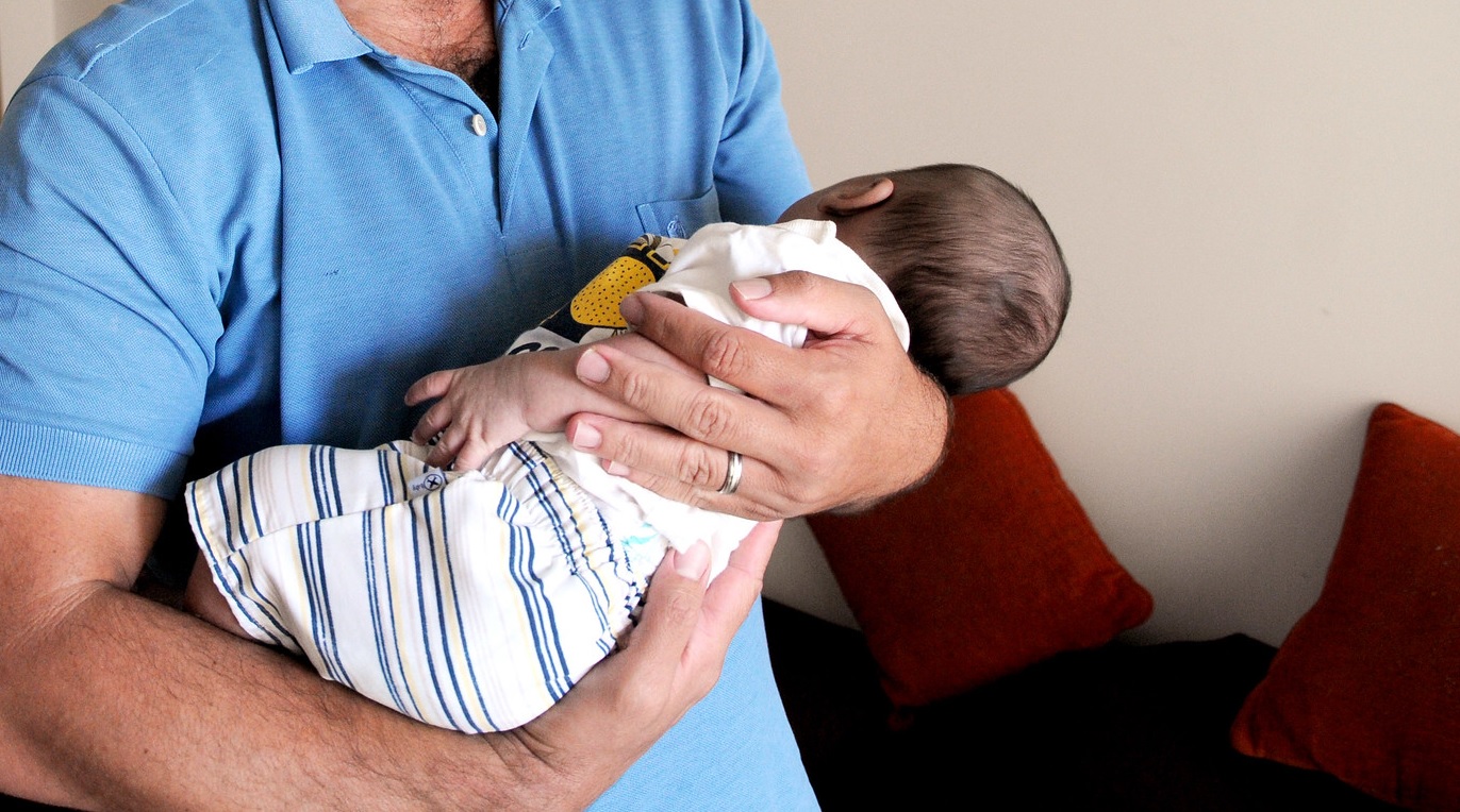 A imagem mostra um adulto vestindo camisa azul segurando um bebê recém-nascido nos braços, com cuidado e proteção. O bebê está de costas, apoiado no peito do adulto.