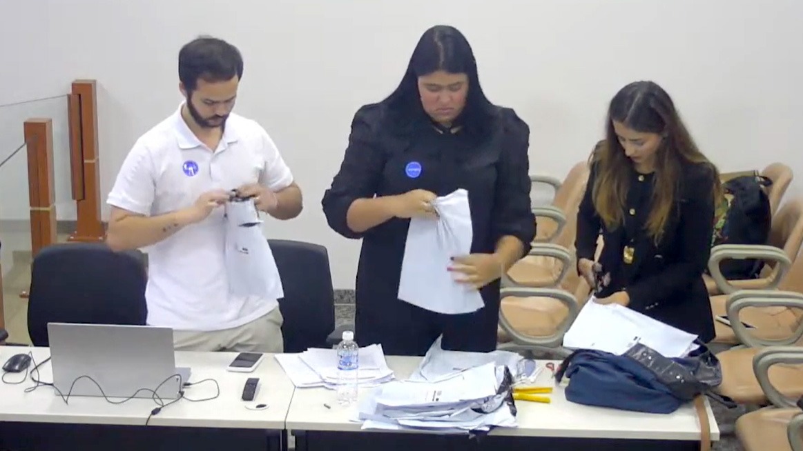 Imagem mostra três pessoas, uma do sexo masculino e duas do feminino, em pé, verificando documentos