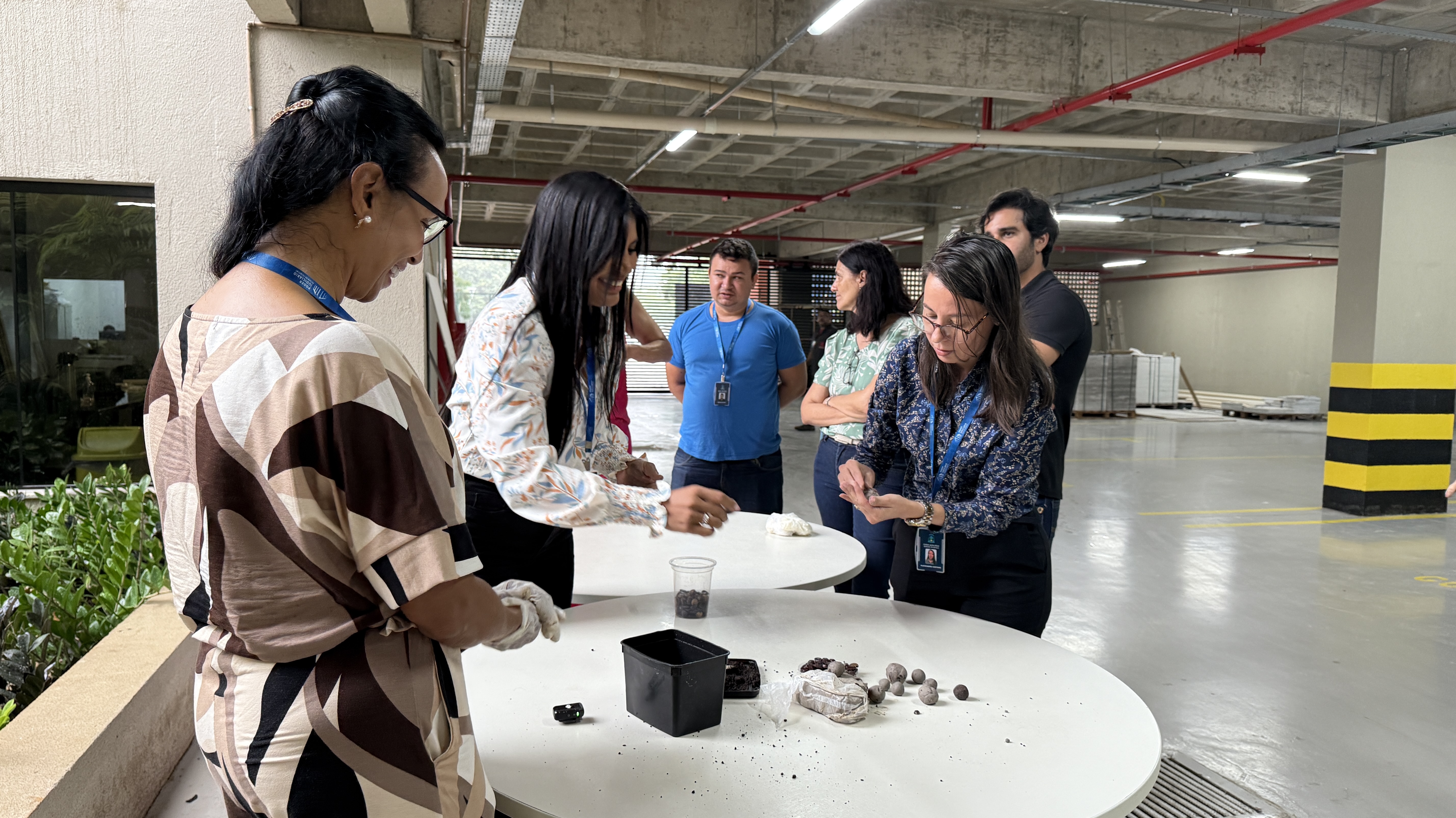 A imagem mostra servidores (as) do Tribunal de Justiça do Tocantins participando da oficina de confecção de bombinhas de sementes, reunidos em torno de uma mesa branca, em um espaço amplo de estacionamento coberto do TJTO. Sobre a mesa, há argila, sementes, recipientes e pequenos materiais utilizados na atividade prática. Em destaque, servidoras moldam as bombinhas com as mãos, enquanto outros participantes observam atentamente o processo.