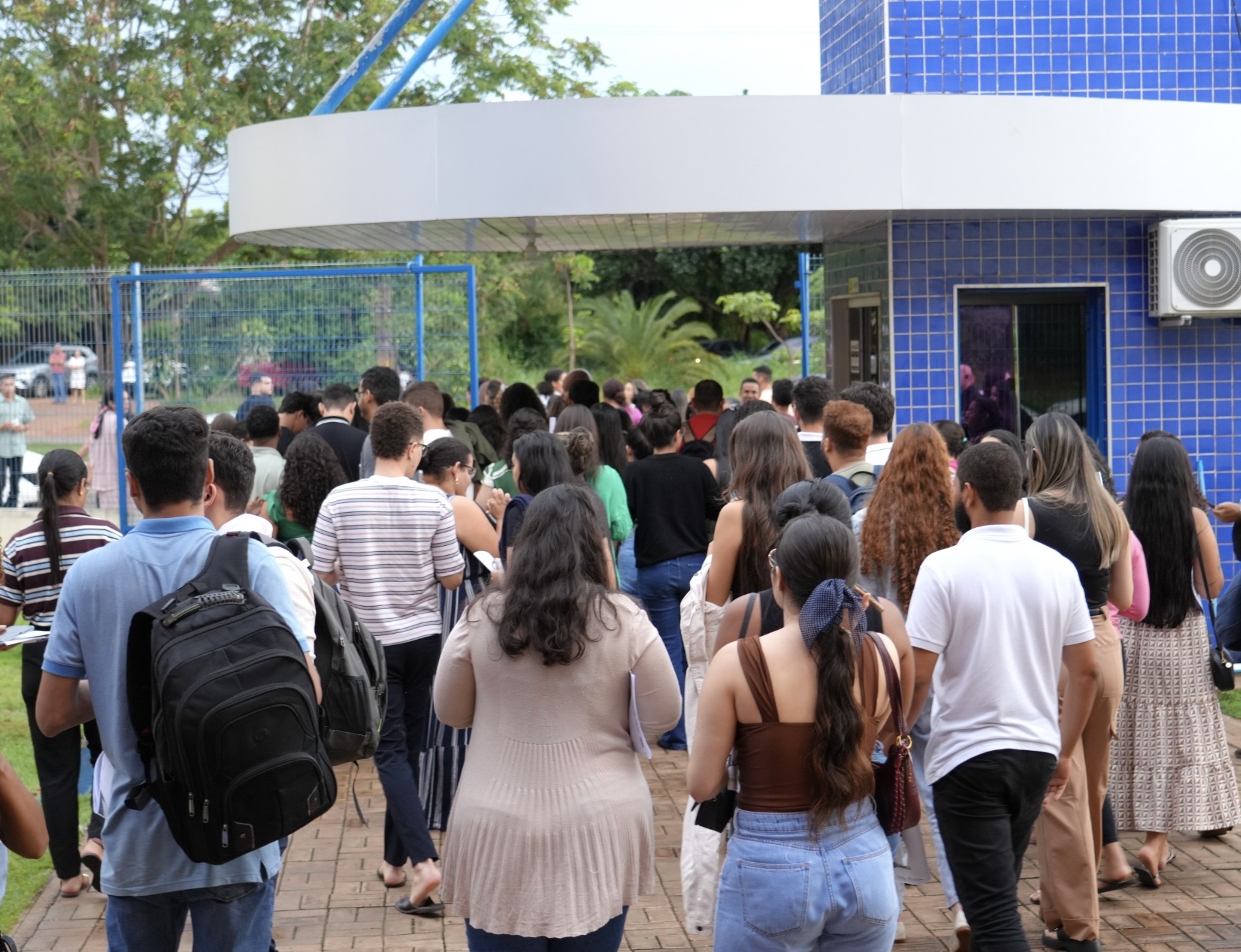 A imagem mostra um grande grupo de pessoas caminhando em direção à entrada de um prédio, para fazer a prova do Programa de Residência com Acesso a Pós-Graduação em Prática Judiciária (PRJud). O local possui portão e estrutura com revestimento azul, enquanto os participantes seguem juntos pelo caminho externo do espaço.