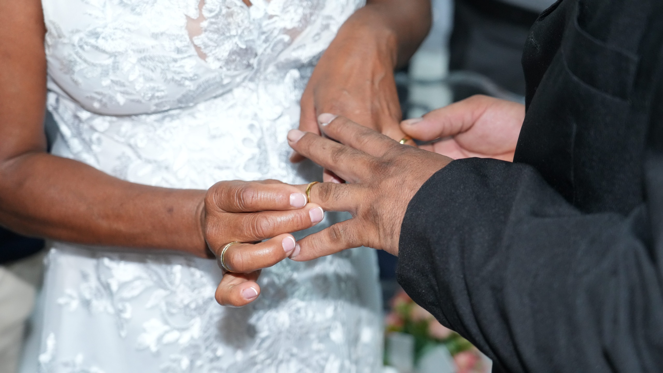 Close nas mãos de um casal durante a cerimônia de casamento. A noiva, vestindo um vestido branco com renda, coloca a aliança no dedo do noivo, que usa traje escuro. O momento simboliza a troca de alianças e a oficialização da união