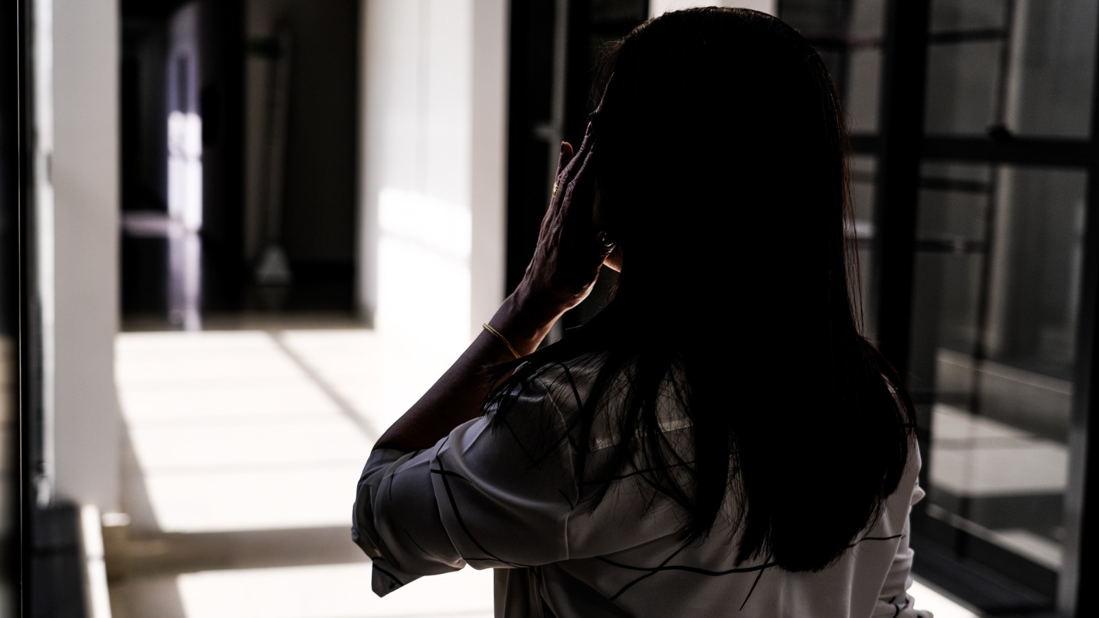 Mulher de costas em corredor interno, segurando celular no ouvido. Ambiente com colunas brancas, paredes de vidro e luz intensa ao fundo.