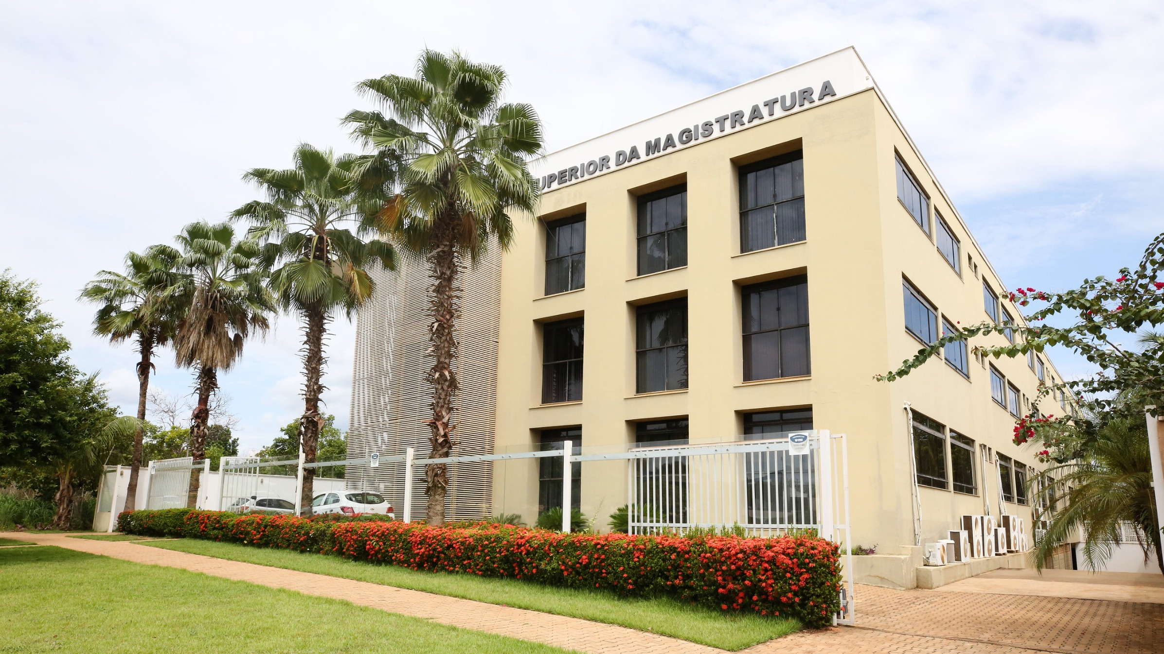 Fachada da Escola Superior da Magistratura, prédio moderno de três andares, em tom bege, com janelas amplas e o nome da instituição no topo. À frente, há jardim com gramado, flores vermelhas e palmeiras, além de grade branca que delimita o espaço.