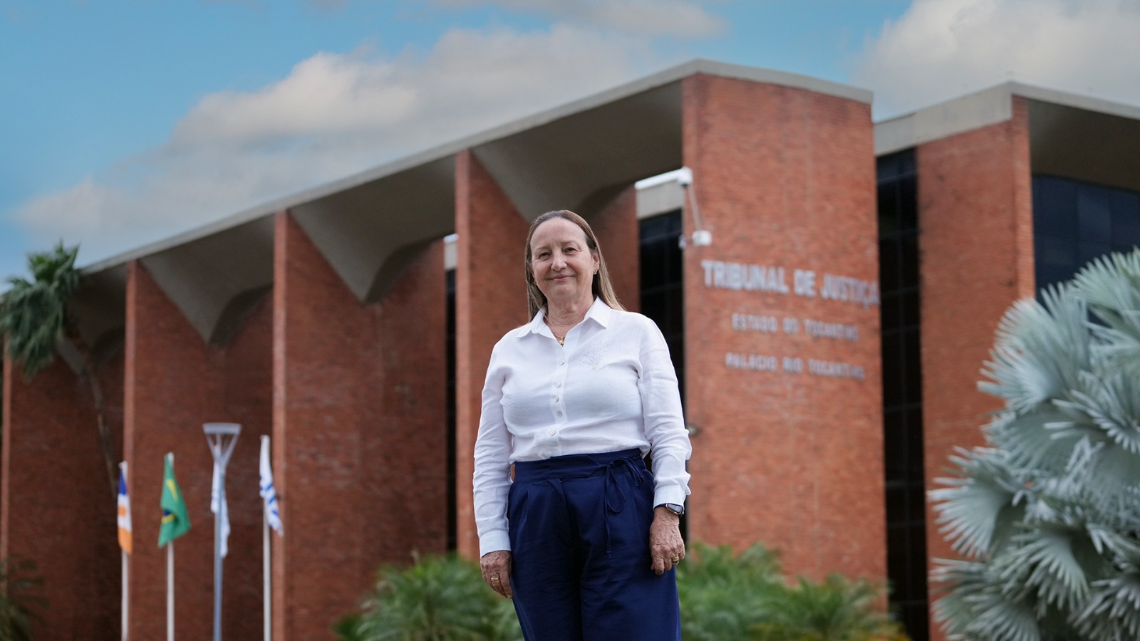 A imagem mostra a presidente do TJTO, desembargadora Maysa Vendramini Rosal, posicionada em primeiro plano, em frente ao prédio do Tribunal de Justiça do Estado do Tocantins. Ela está em pé, olhando para a câmera, com postura ereta e expressão serena. Veste camisa branca de manga longa e calça social azul-marinho de cintura alta, compondo um visual formal e institucional. Ao fundo aparece a fachada do prédio do tribunal, construída em tijolos aparentes, com estrutura moderna marcada por grandes pilares e cobertura em linhas geométricas. Na parede é possível ler a identificação do local: “Tribunal de Justiça – Estado do Tocantins – Palácio Rio Tocantins”.
